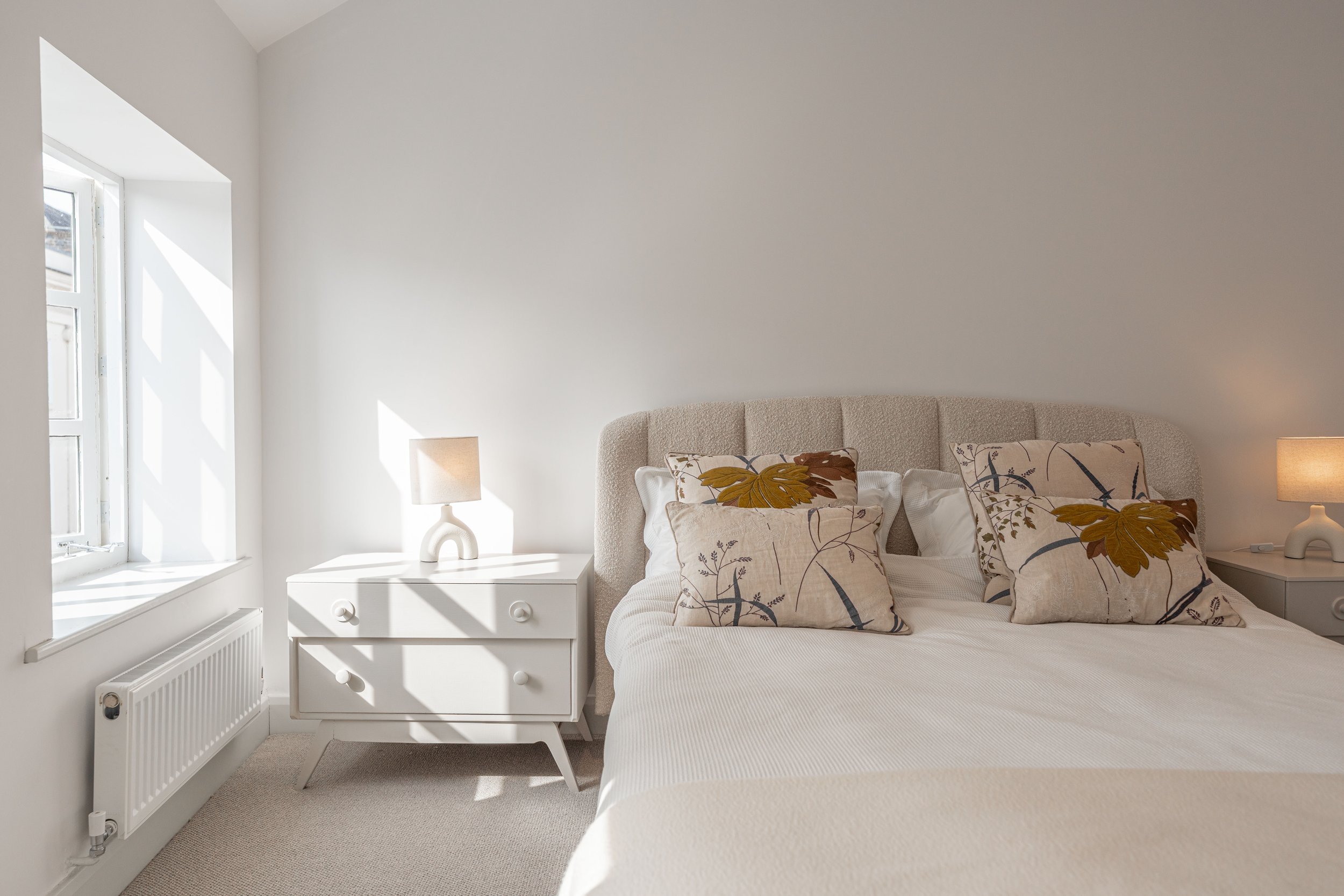 A neatly made bed with four pillows featuring floral designs in shades of brown, yellow, and gray, positioned against a beige upholstered headboard in a bright, minimalist bedroom with white walls and a window.