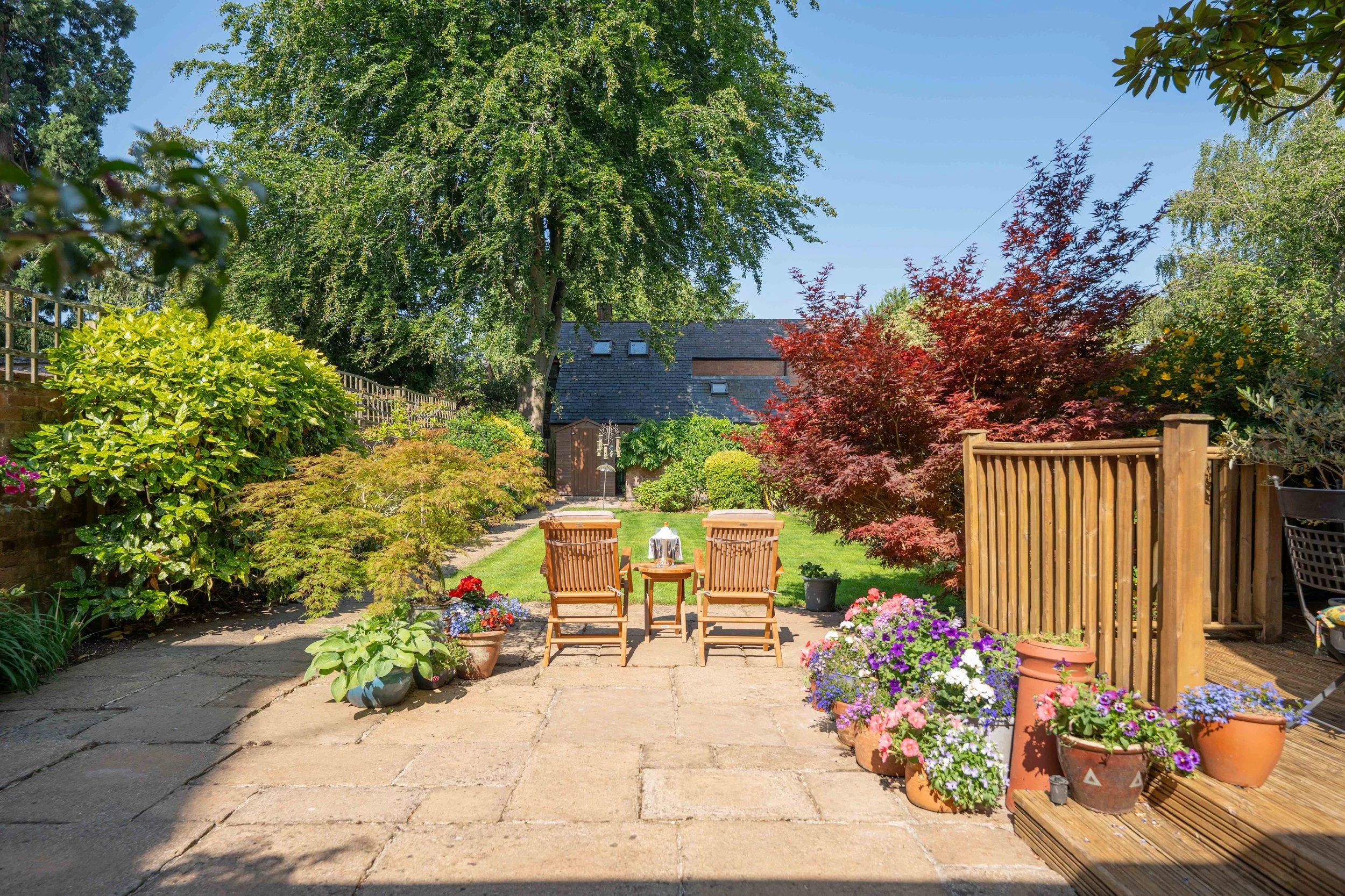 A backyard patio with two wooden chairs and a small table, surrounded by colorful potted flowers, green bushes, a tree with red leaves, and a lawn leading to a brick house with a slate roof and skylights.