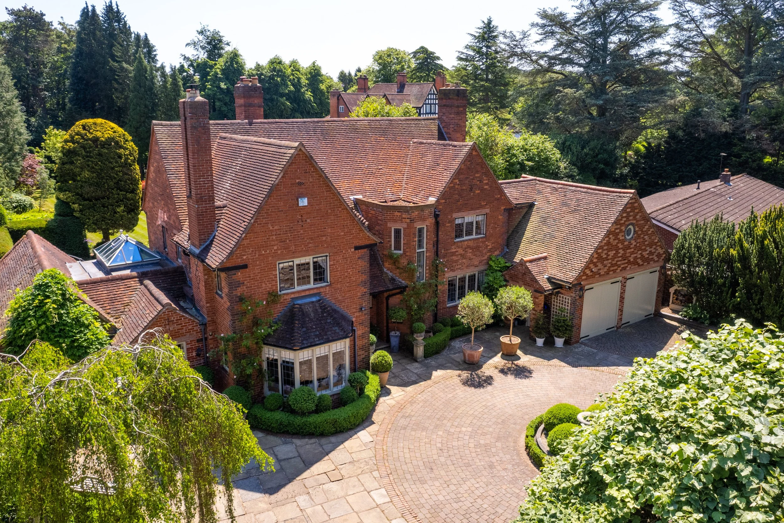 Aerial view of a large brick house with landscaped front yard, potted plants, trees, and adjacent garage in a lush suburban neighborhood.