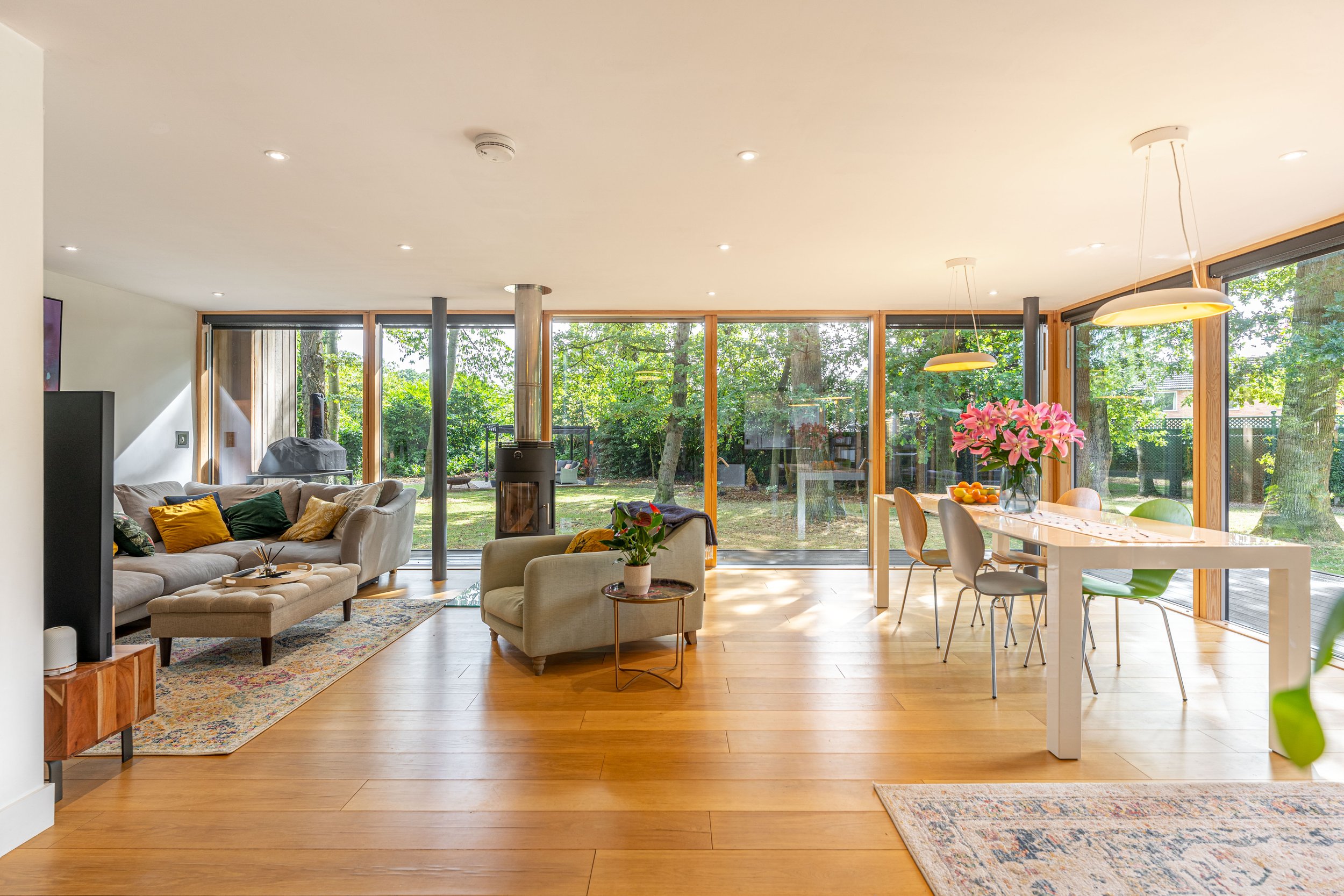 Bright open living and dining area with large floor-to-ceiling windows showing a garden outside. The space includes a beige sofa with pillows, a small armchair, a floral area rug, a modern wood stove, and a white dining table with pink flowers as a c