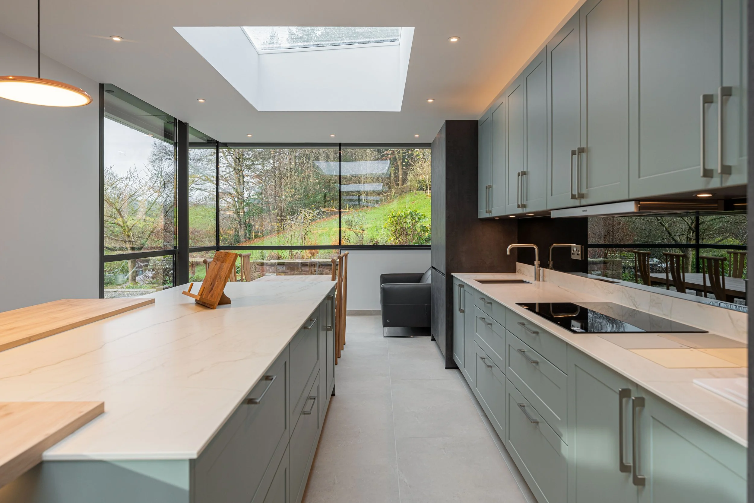 Modern kitchen with light green cabinets, white marble countertops, and large windows revealing a lush outdoor landscape.