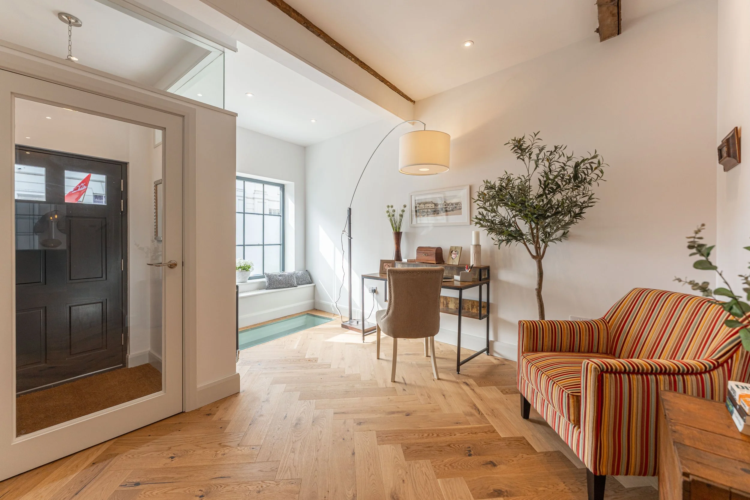 Bright interior with wooden flooring, a striped armchair, a desk with a chair, a large potted plant, a window nook with cushions, and a door with a glass panel leading outside.