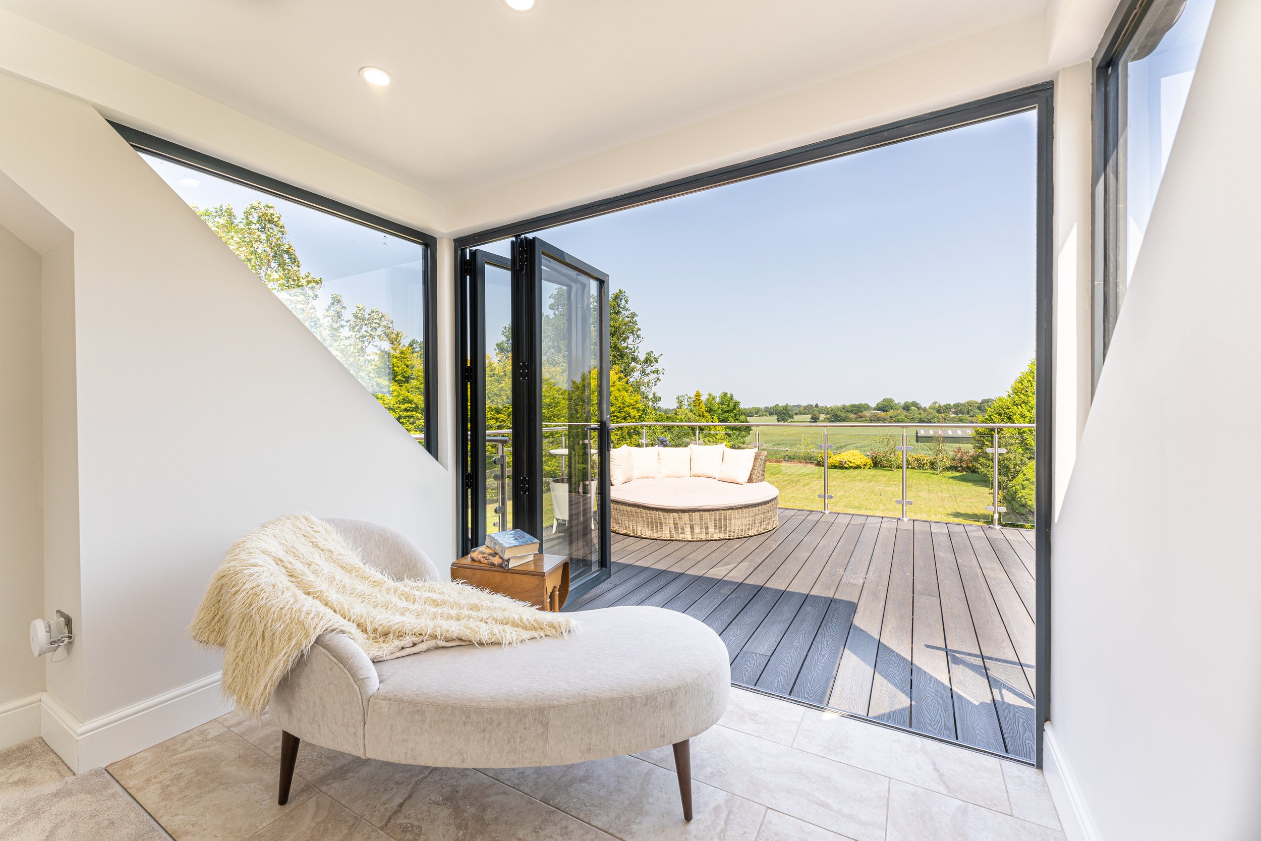 Living room with opened glass doors leading to a wooden balcony with outdoor seating, overlooking a green landscape.