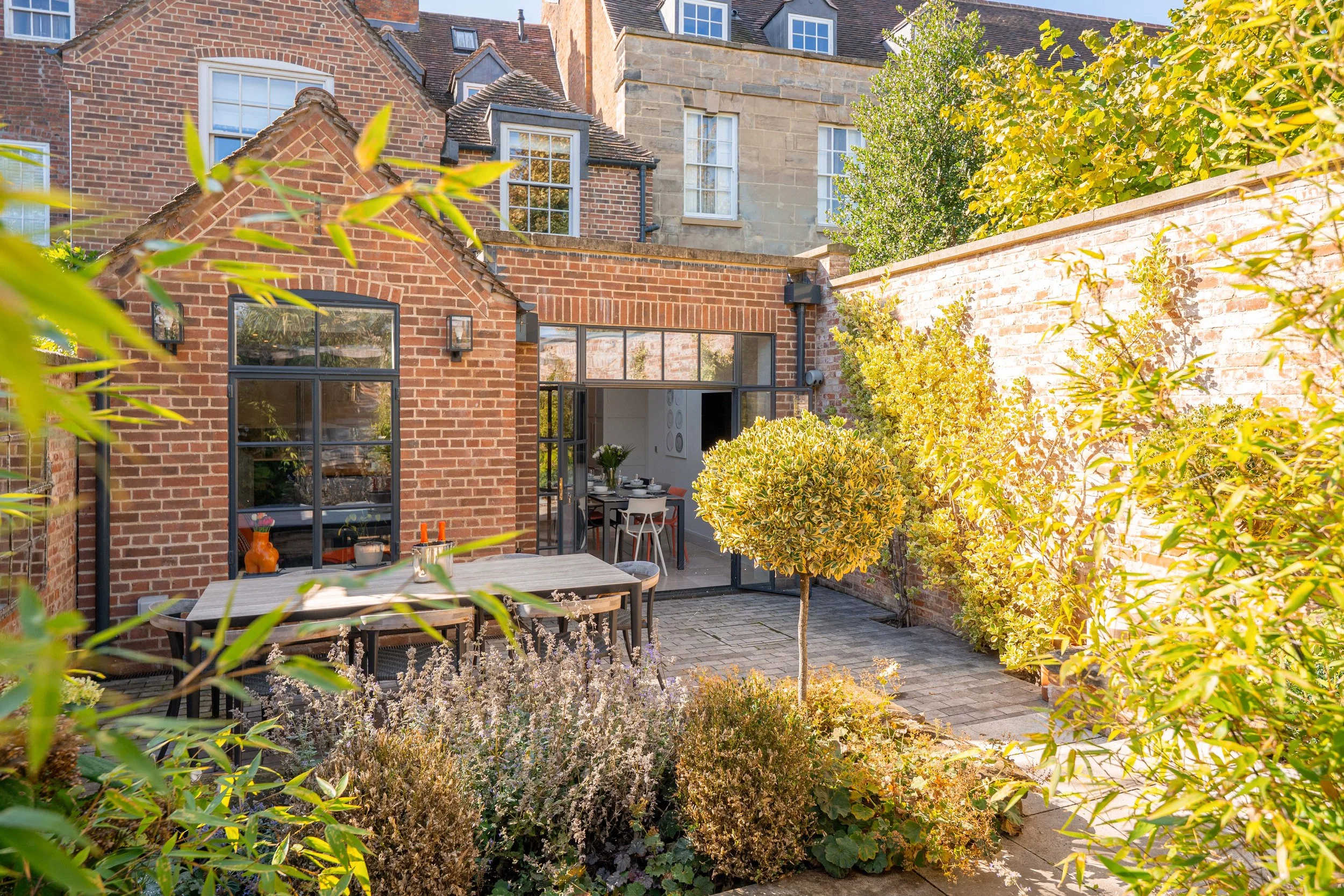 A cozy backyard with brick walls, lush green plants, and patio furniture, overlooking neighboring houses with brick and stone facades.
