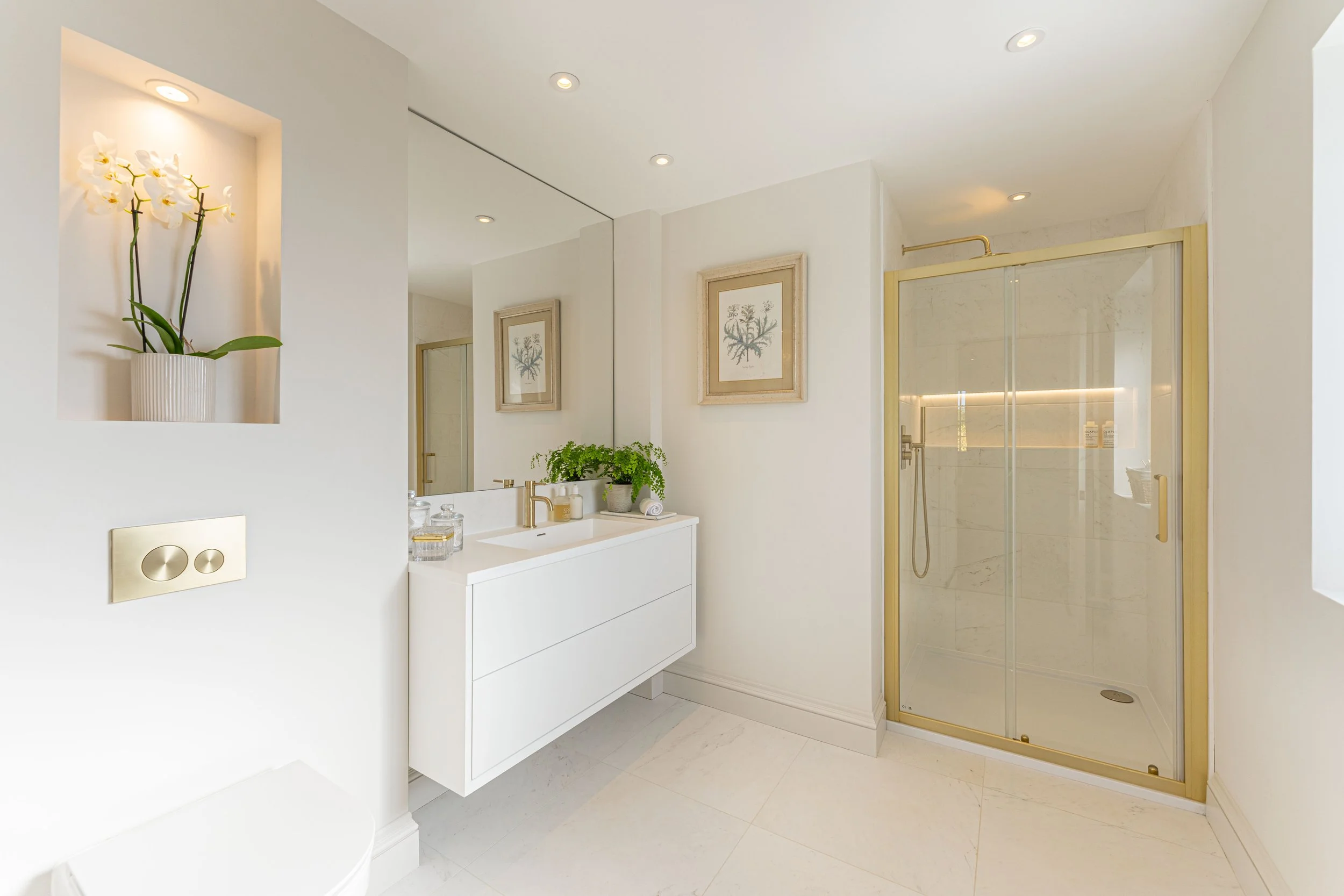 A modern bathroom with a white vanity, large mirror, framed botanical wall art, potted green plant, and a glass-enclosed shower with gold fixtures.