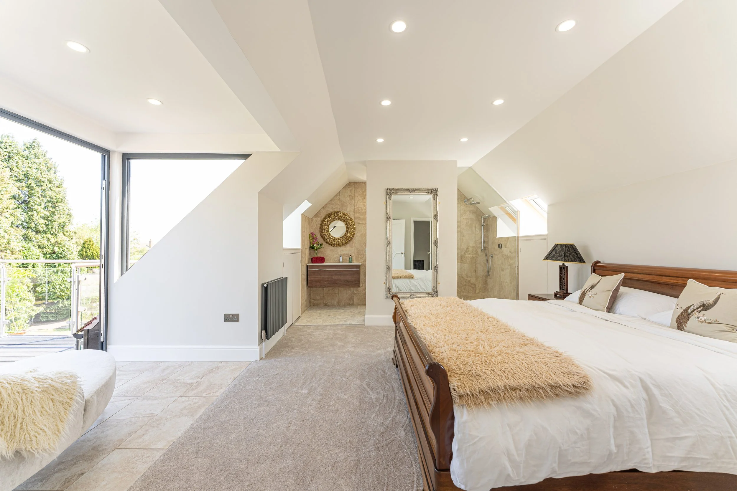 A modern bedroom with a large bed, decorative pillows, a side table with a lamp, an ornate mirror, and a walk-in shower with natural light coming through skylights.