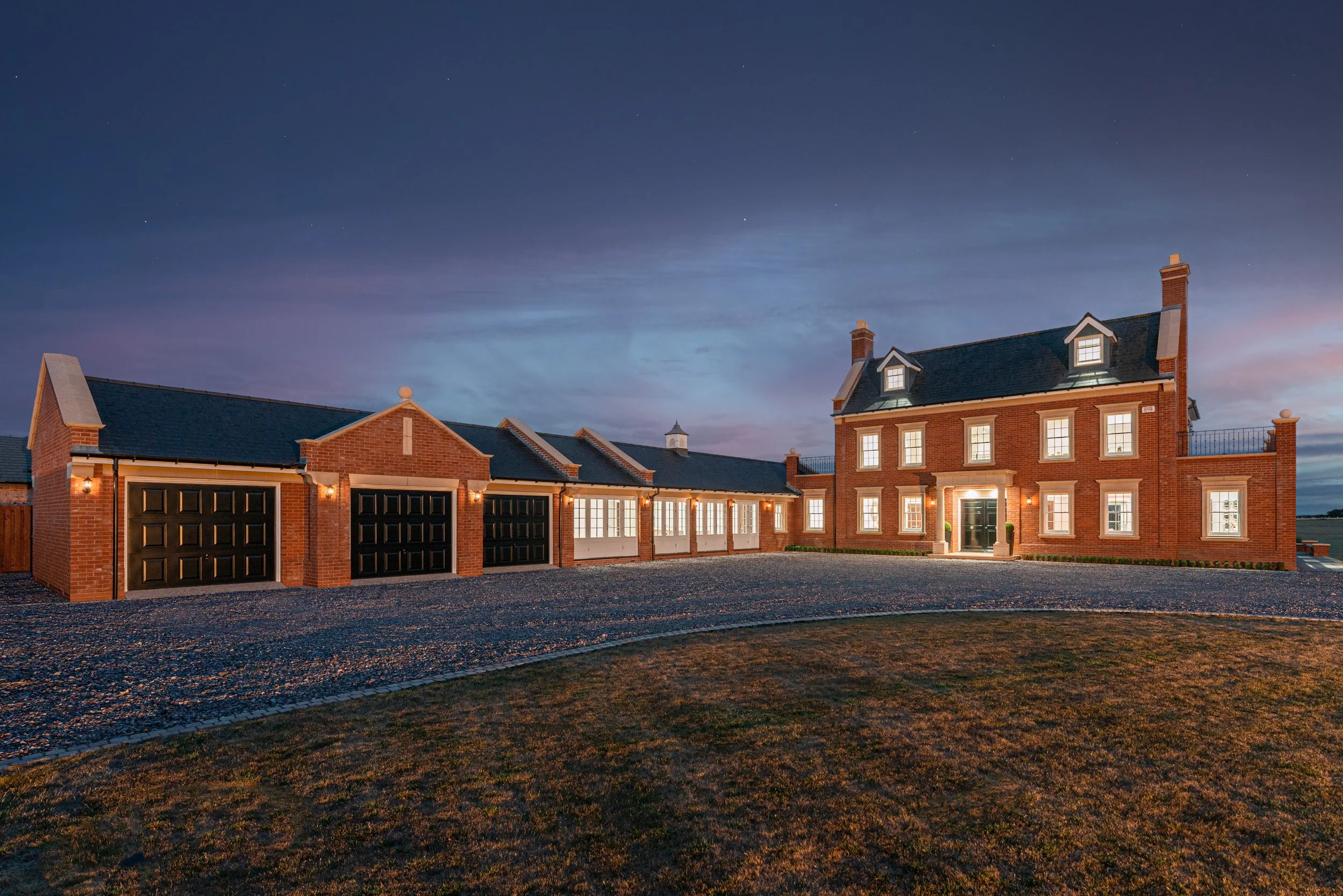 A large brick house with multiple windows and a dark roof, illuminated from inside, during twilight, with garages on the left and a gravel driveway in front.