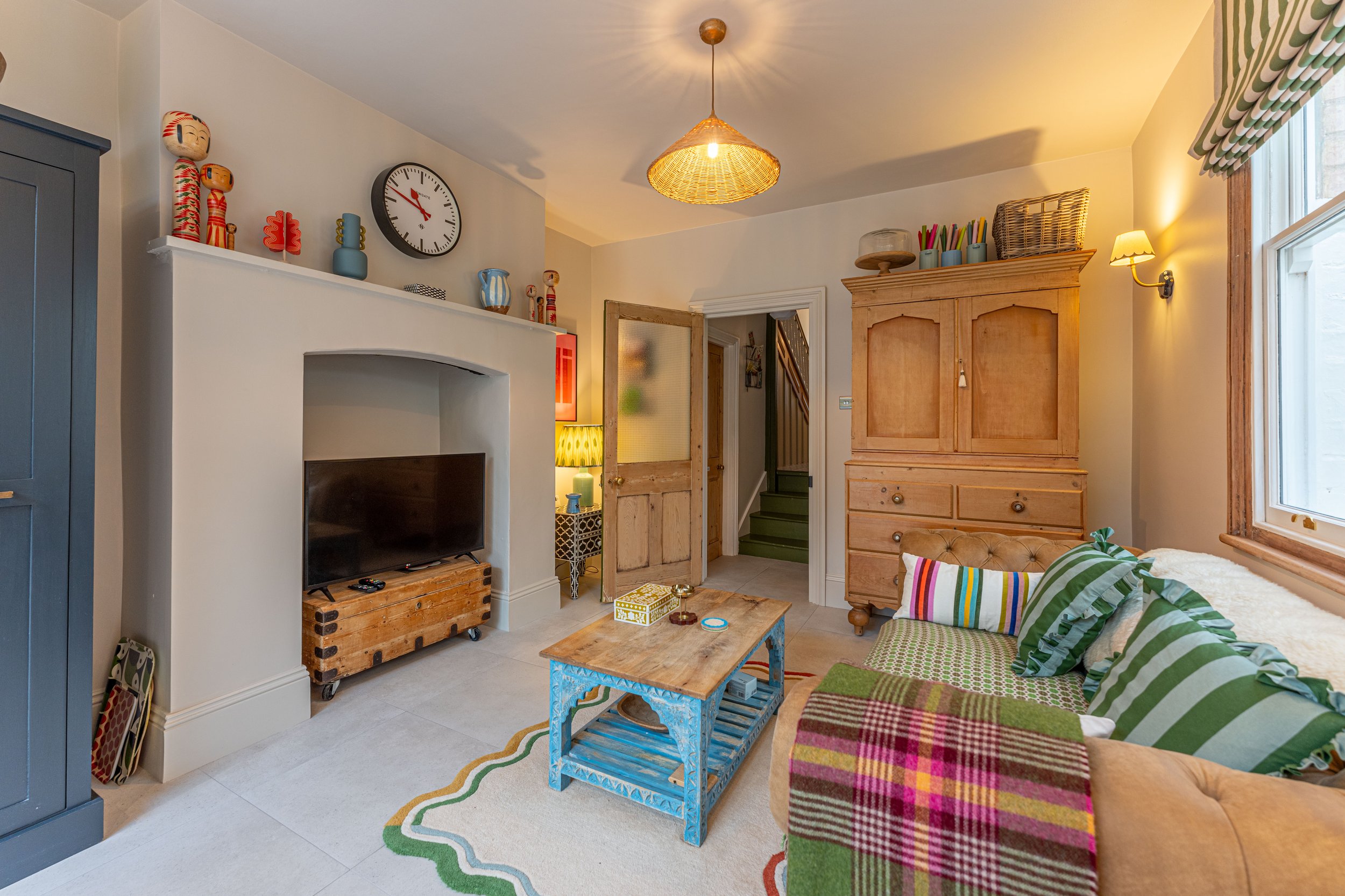 Living room with a beige couch, colorful striped pillows, a wooden coffee table, a TV on a wooden stand, a white wall with shelves holding decorative items, a large wooden wardrobe, a window with striped curtains, a textured ceiling light, and variou