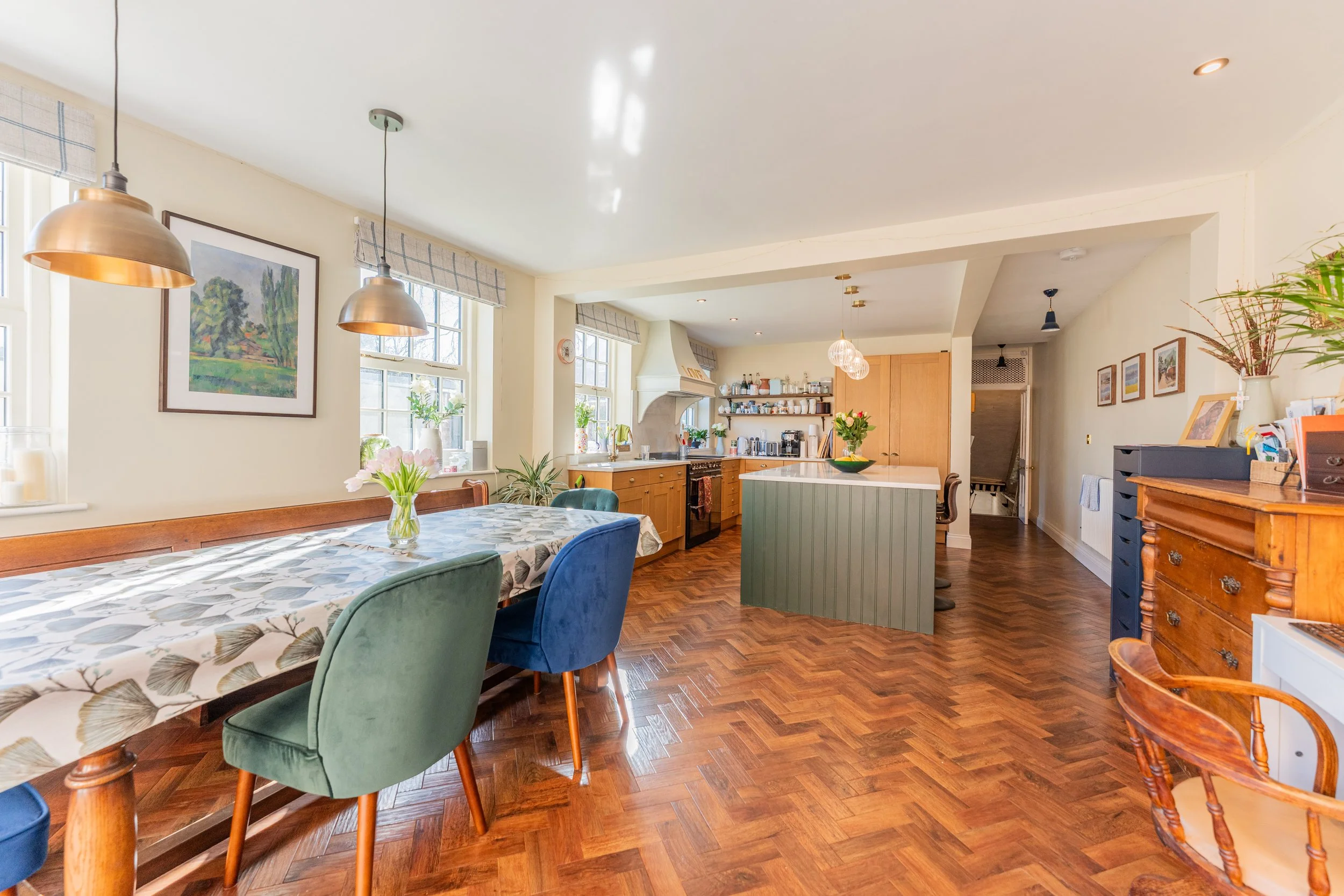 Bright open-concept kitchen and dining area with hardwood floors, a long dining table with colorful chairs, an island with a green panel, pendant lights, and windows with plaid valances.