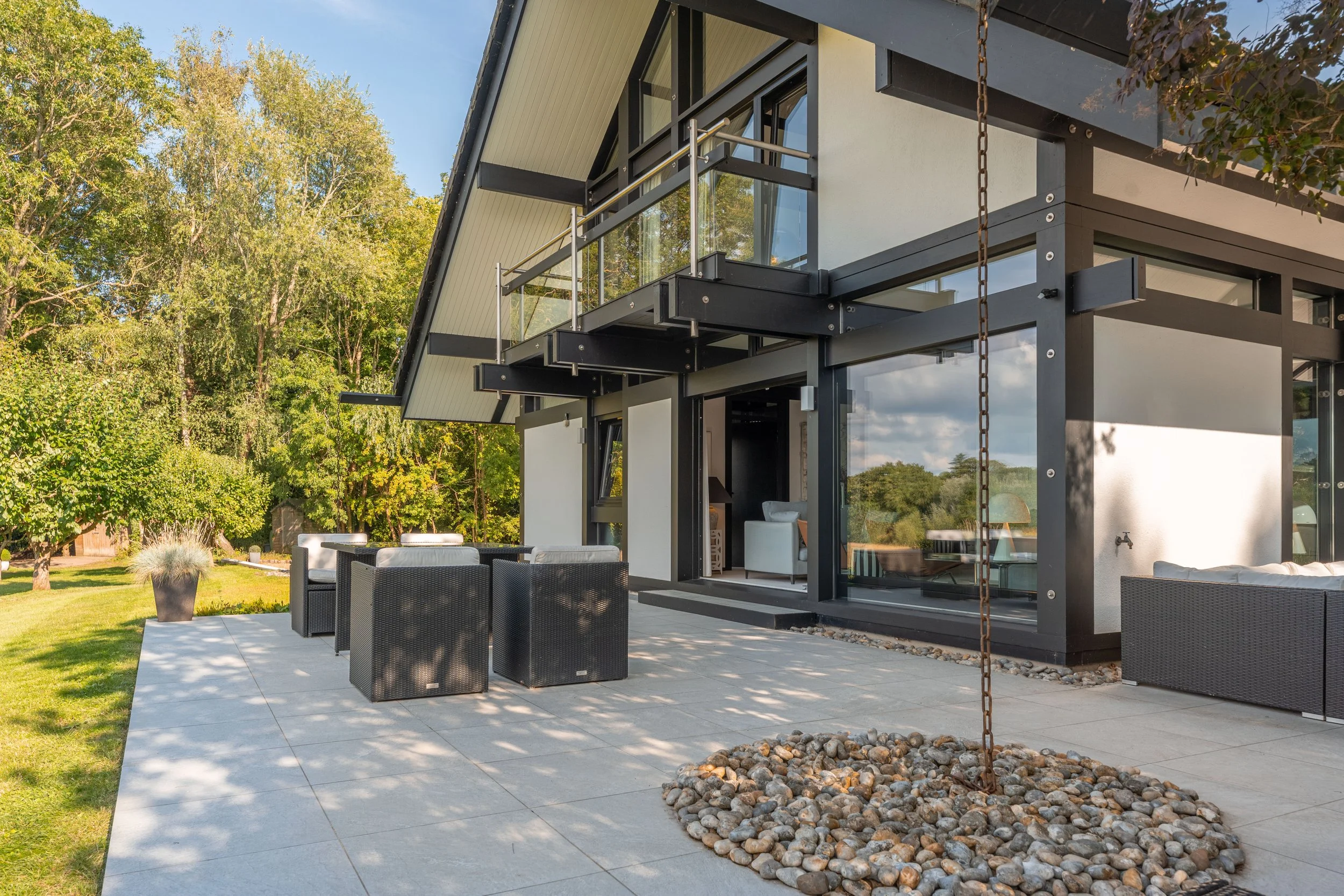 Modern house with large patio seating area, glass railing balcony, and surrounding greenery.