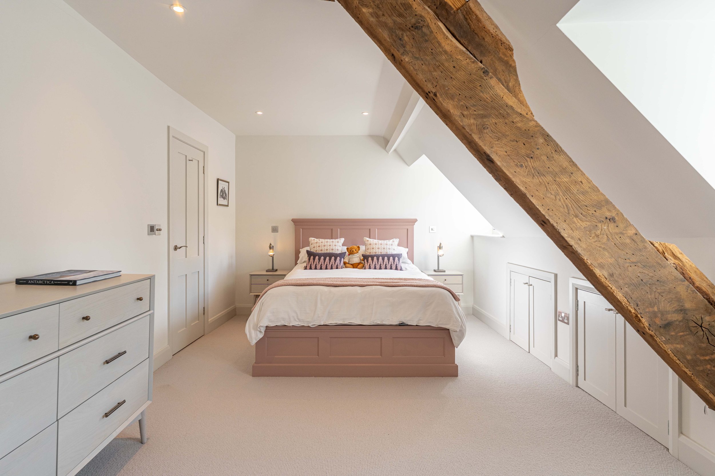 A bedroom with white walls, a pink bed frame, two nightstands with lamps, and a teddy bear on the bed. There's a wooden beam on the ceiling and built-in storage on the right wall.