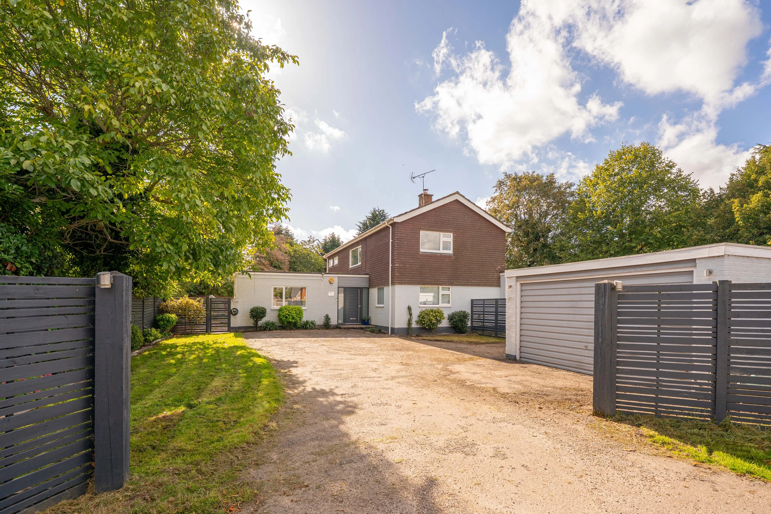 View of a house with a gravel driveway, enclosed by dark grey wooden fences, trees on the left and right, and a partly cloudy sky above.