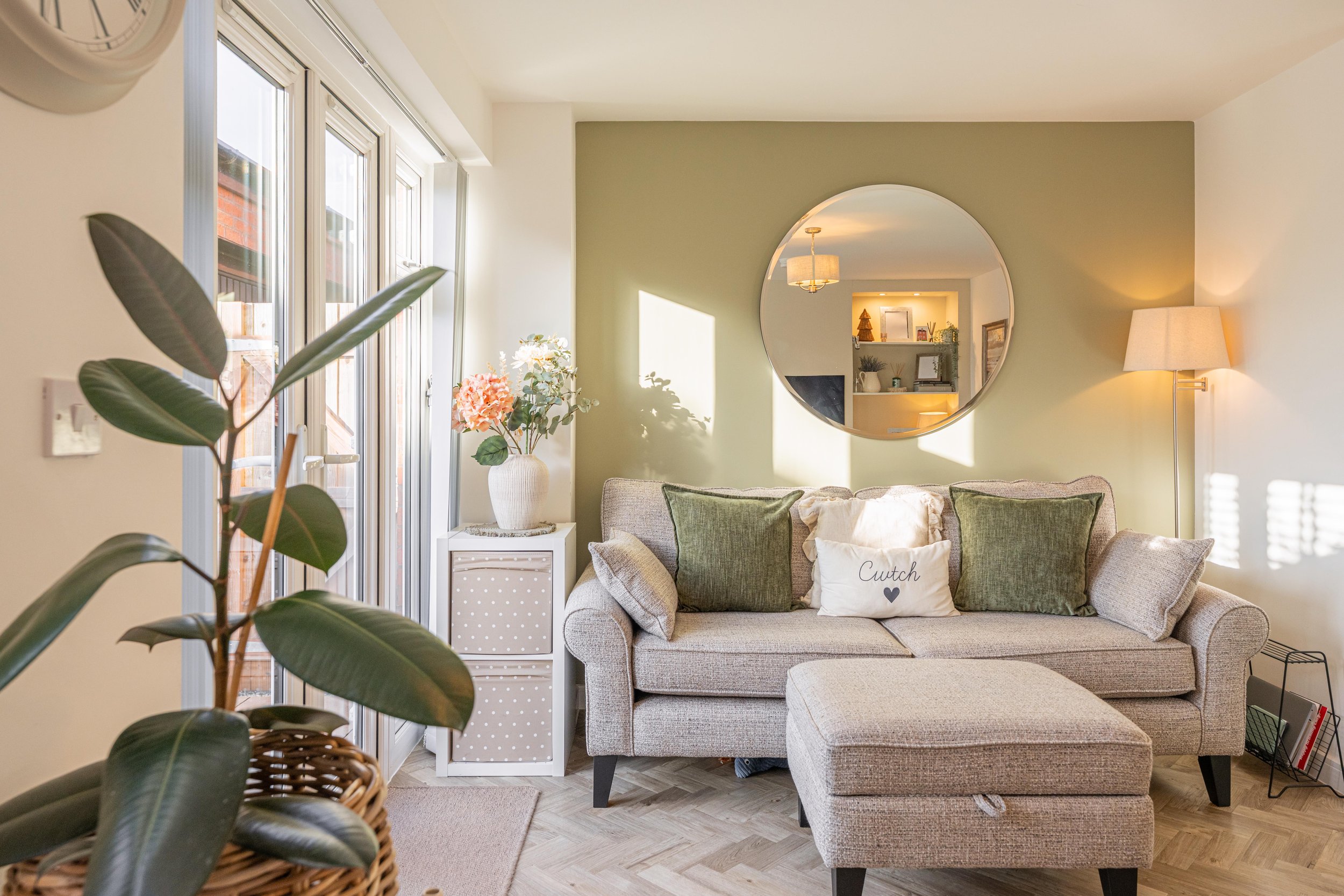 Sunlit living room with a grey sofa, green and white pillows, a round mirror on a light green wall, a tall floor lamp, a white side table with a vase of flowers, large window with blinds, and a magazine rack on the floor.