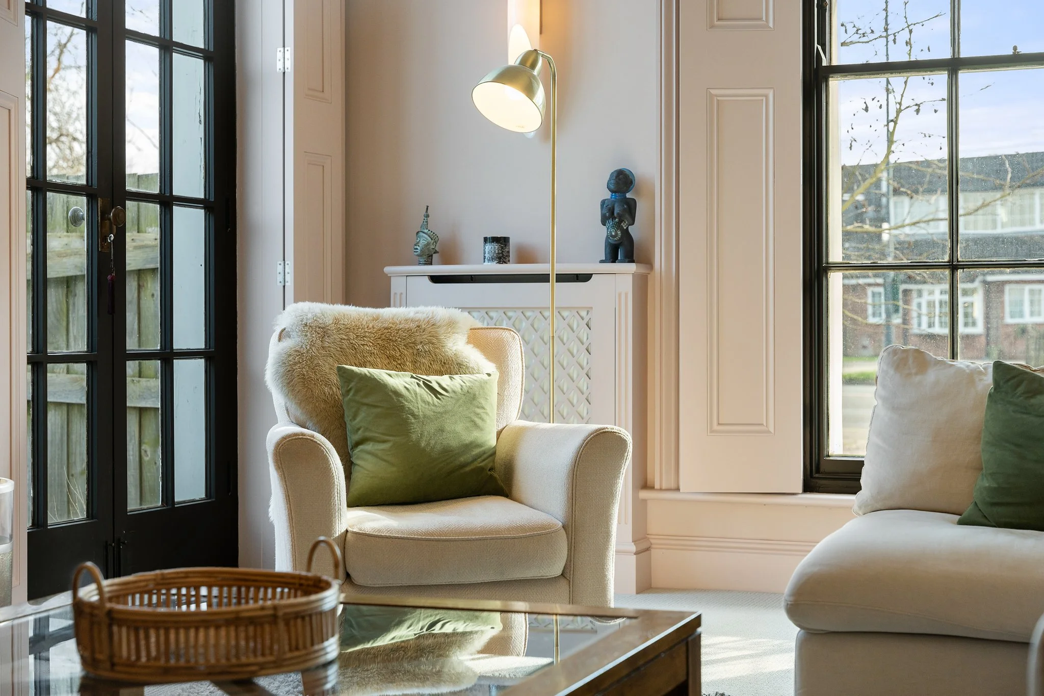 Living room with white armchair, green pillow, cream sofa with green pillow, glass coffee table, black framed windows, and sunlight coming in.