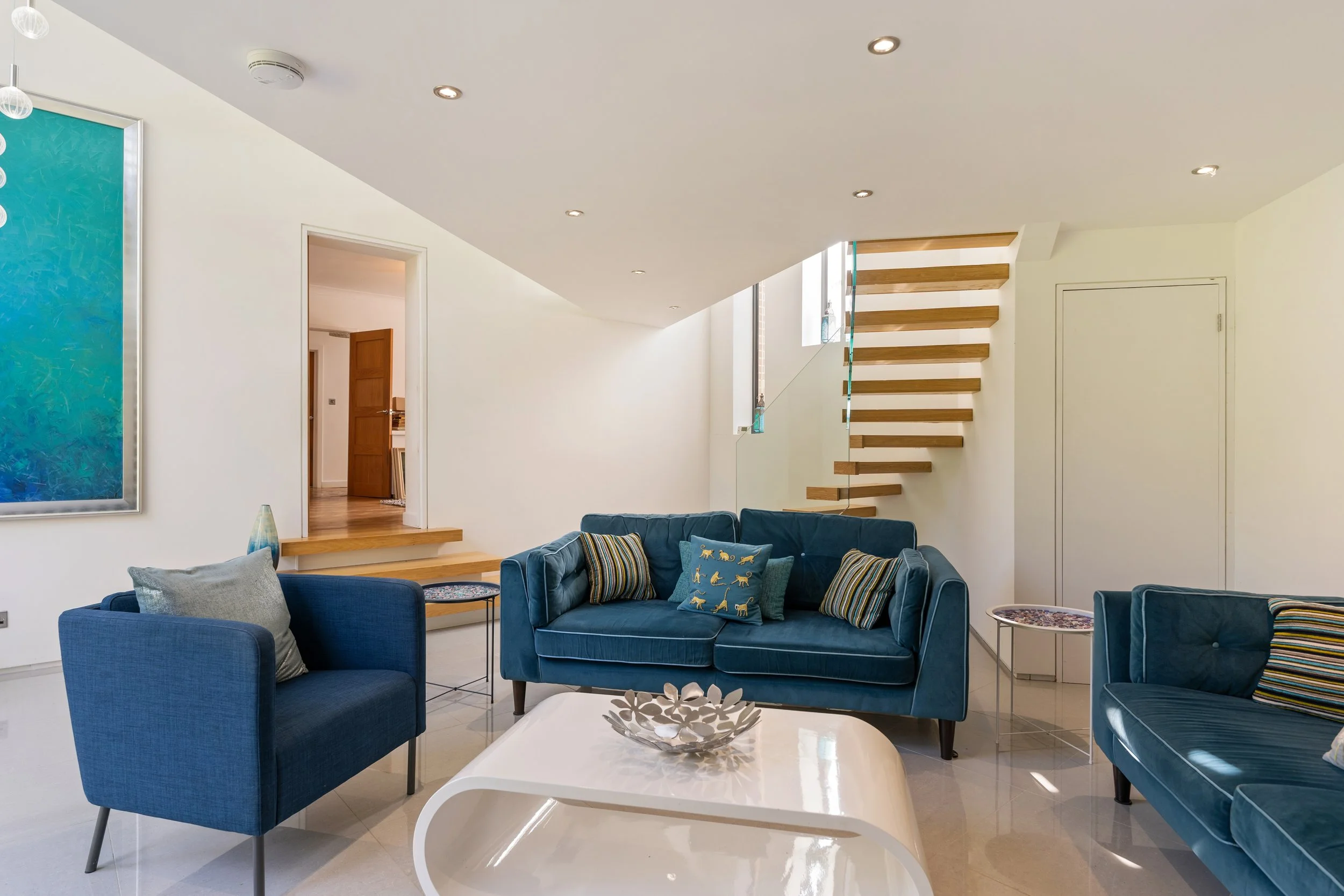 Living room with blue sofas, decorative pillows, a white coffee table, and an open wooden staircase leading to the upper floor, with natural light coming from windows.