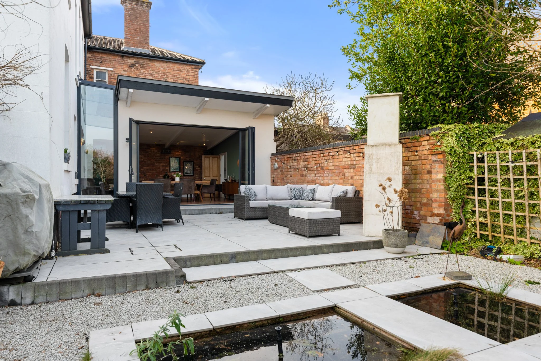 Backyard patio with outdoor seating, plastic patio furniture, a wicker sectional sofa with cushions, and a small pond with a black reflective water surface, surrounded by white paving stones and gravel, enclosed by brick and concrete walls, with tree
