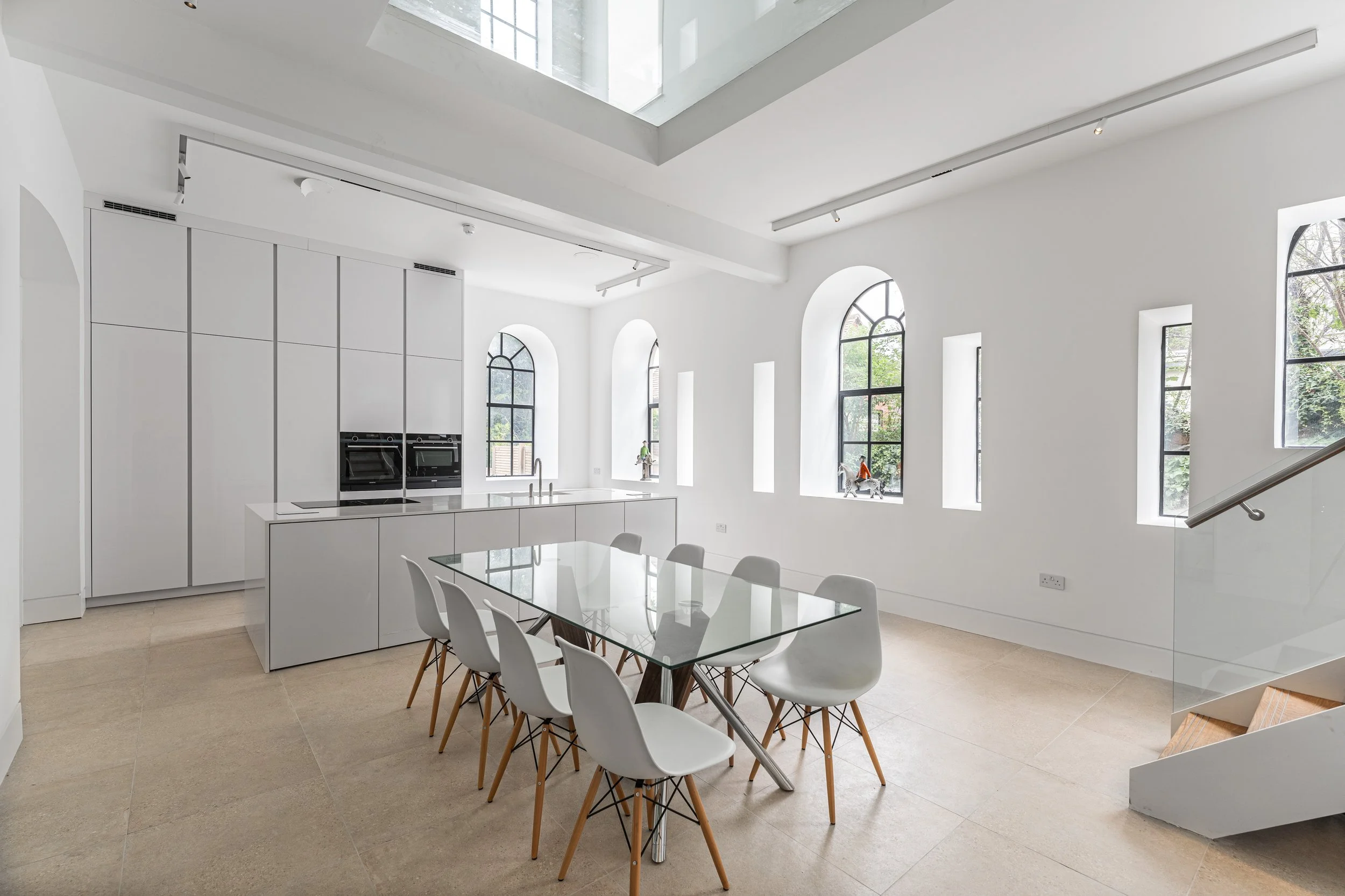 Modern, minimalistic dining area with a glass-top table and white chairs, large arched windows with black frames, a staircase with a glass railing, and a white kitchen with hidden appliances.