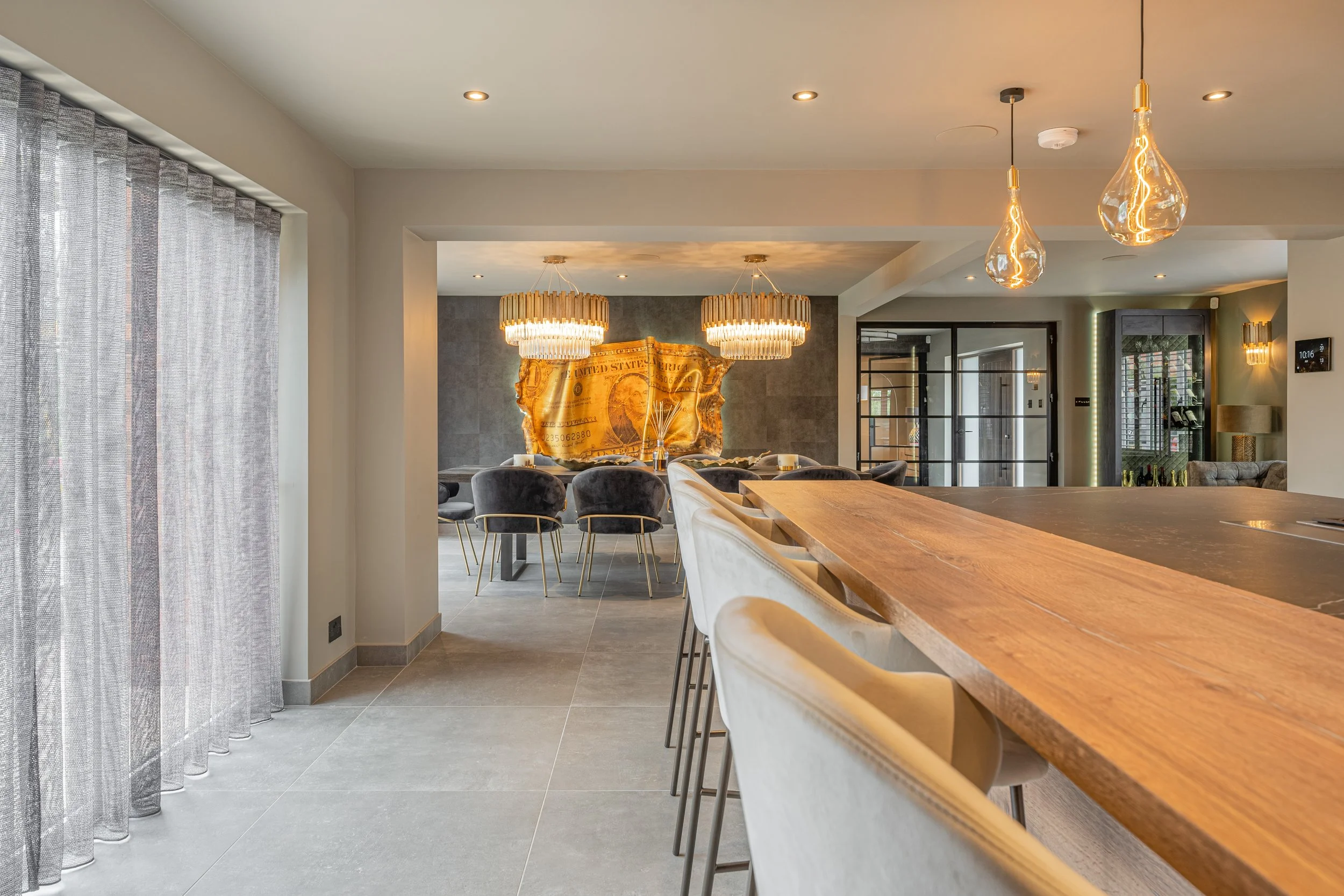 Modern dining and kitchen area with pendant lights, a large wooden island, velvet chairs, a wall art of a dollar bill, and sliding glass doors