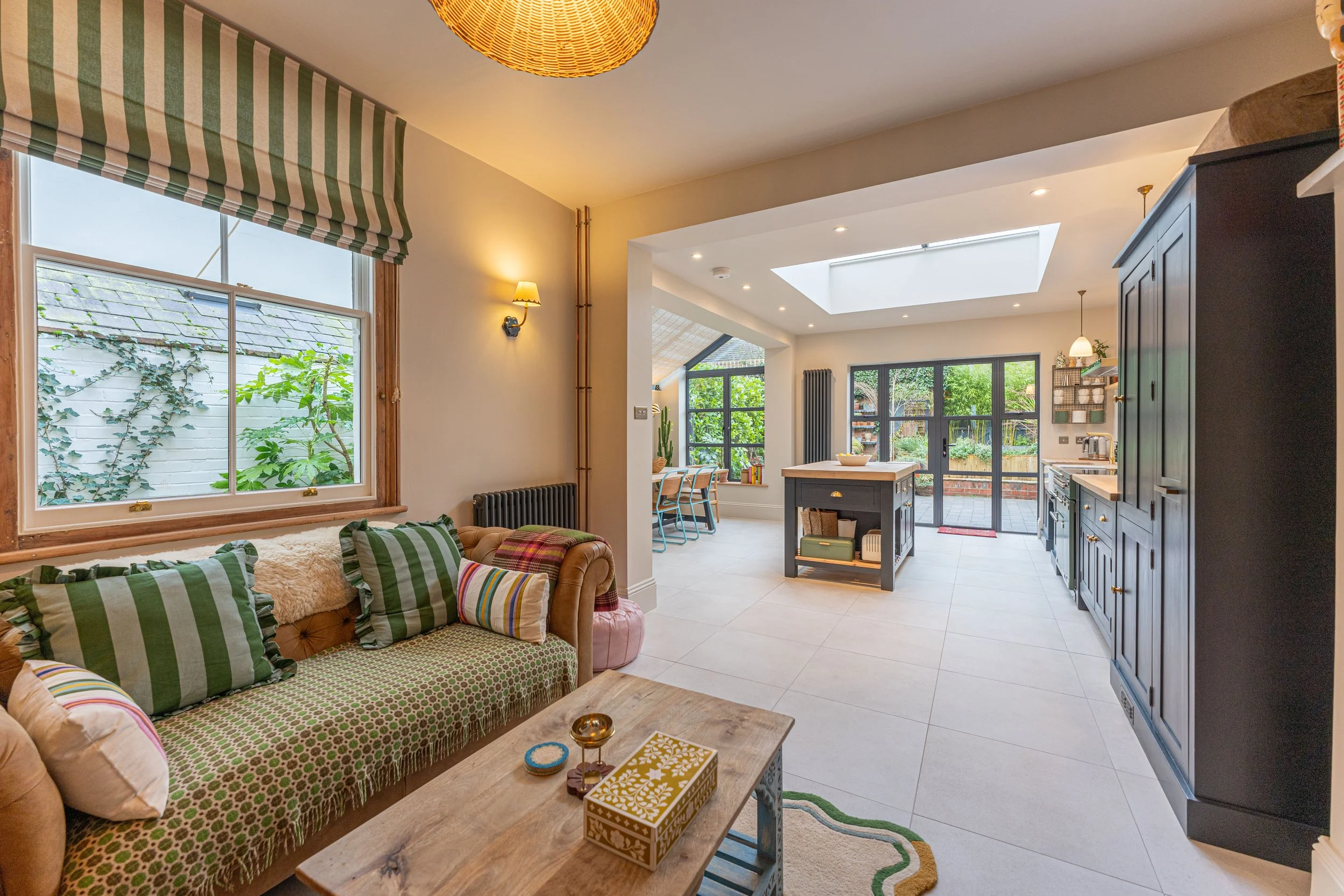 Open-plan living room and kitchen with large window, skylight, and sliding glass doors, featuring a cozy sofa with striped pillows, a rustic wooden coffee table, and modern kitchen cabinets with pendant lights, overlooking a garden.