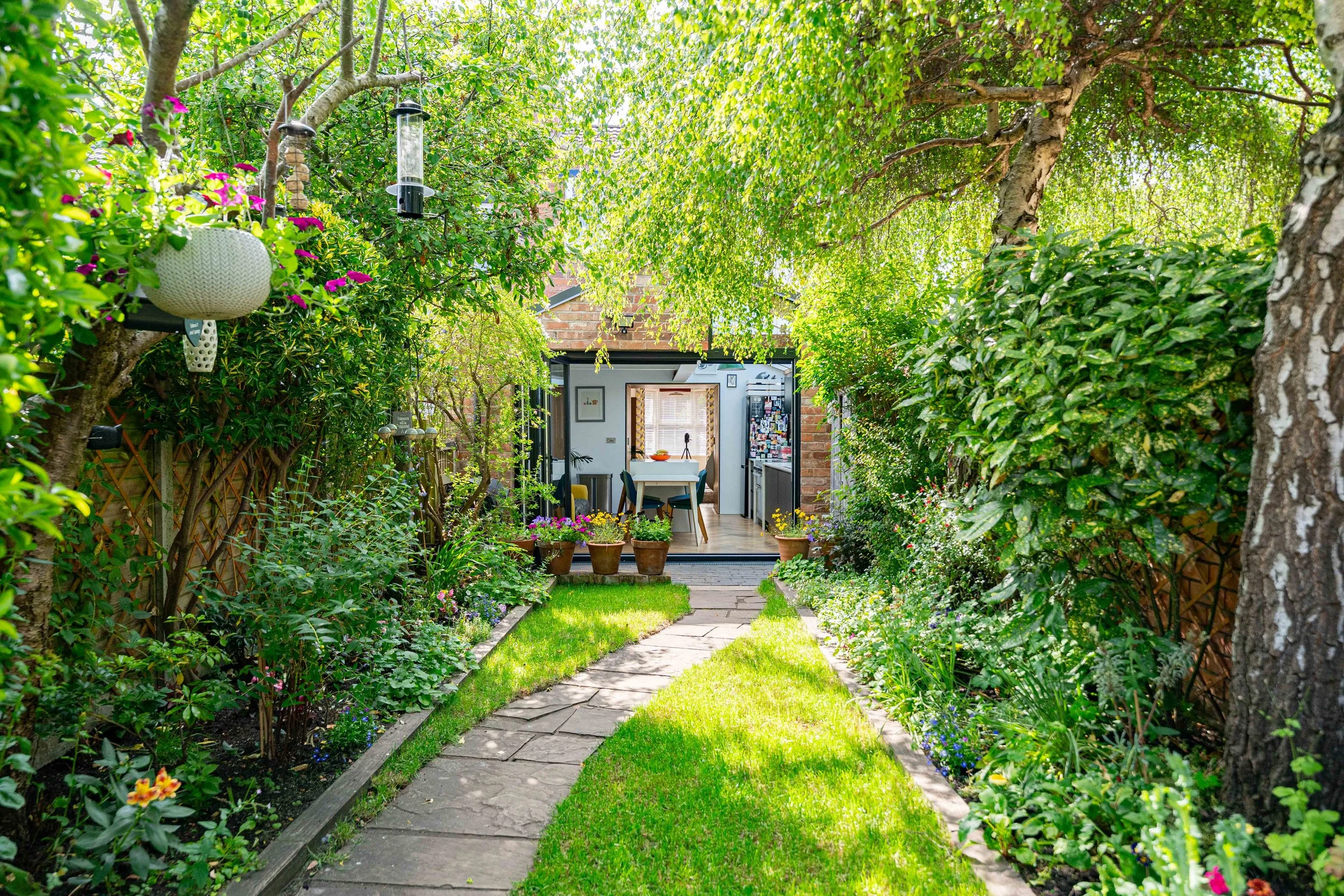 A lush garden path with stone tiles, green grass, and vibrant flowers leading to a cozy outdoor kitchen and dining area under a canopy of trees and shrubs.