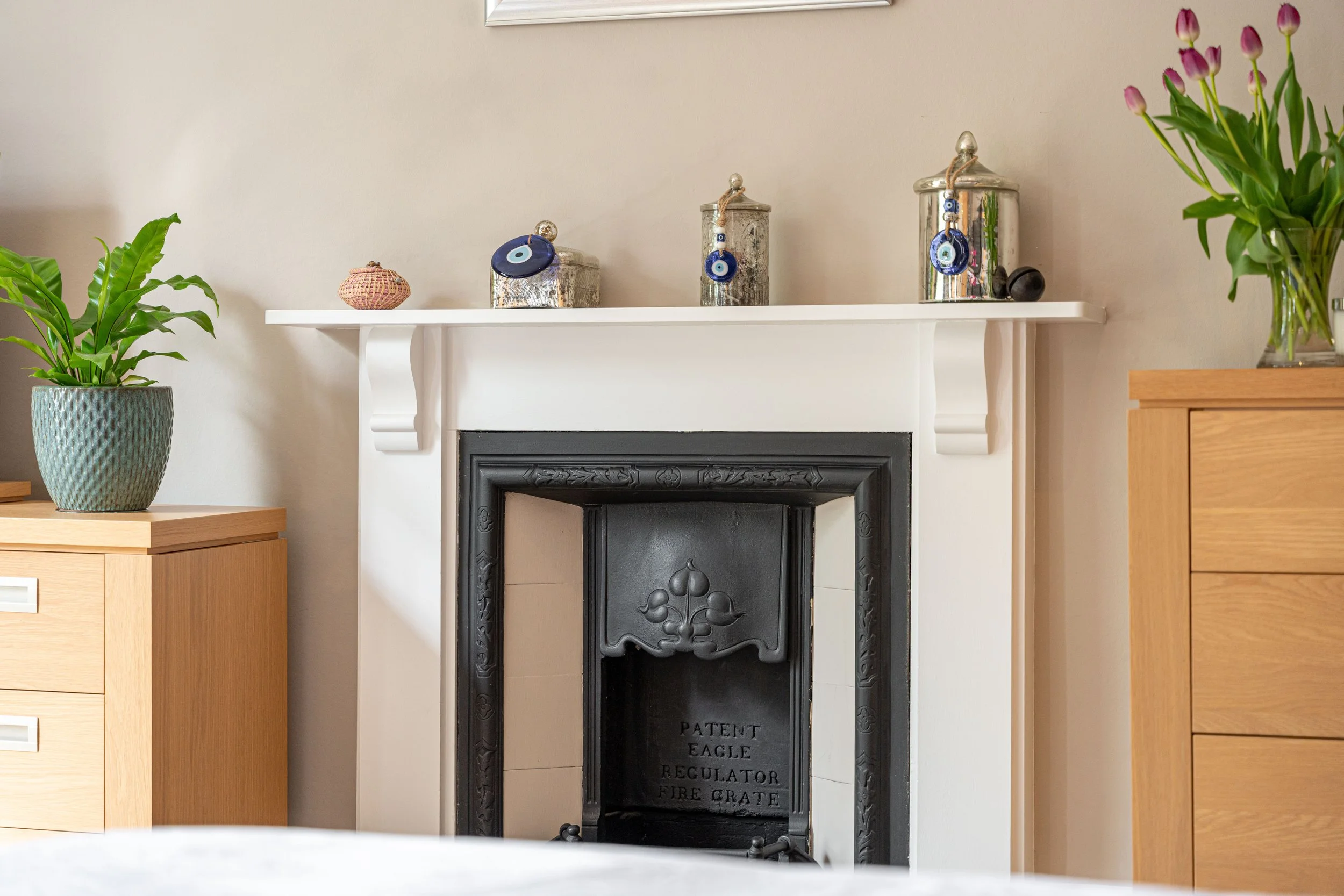 Living room fireplace with mantel decorated with silver containers, small glass figurine, wall art, and vases with pink tulips, and two wooden cabinets with potted plants.