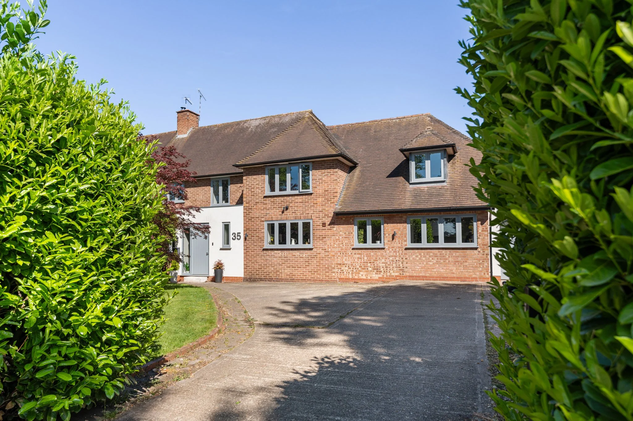 A two-story brick house with a white front door, numbered 35, and multiple windows, surrounded by green hedges and trees under a clear blue sky.