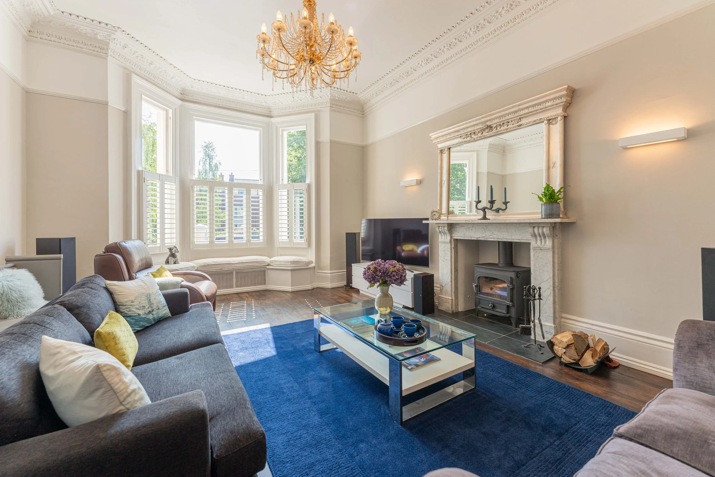 A living room with a gray sofa, a brown armchair, a blue area rug, and a glass coffee table with a vase of purple flowers. There is a television, fireplace with a mirror above, and built-in window seat with cushions near a large bay window with white