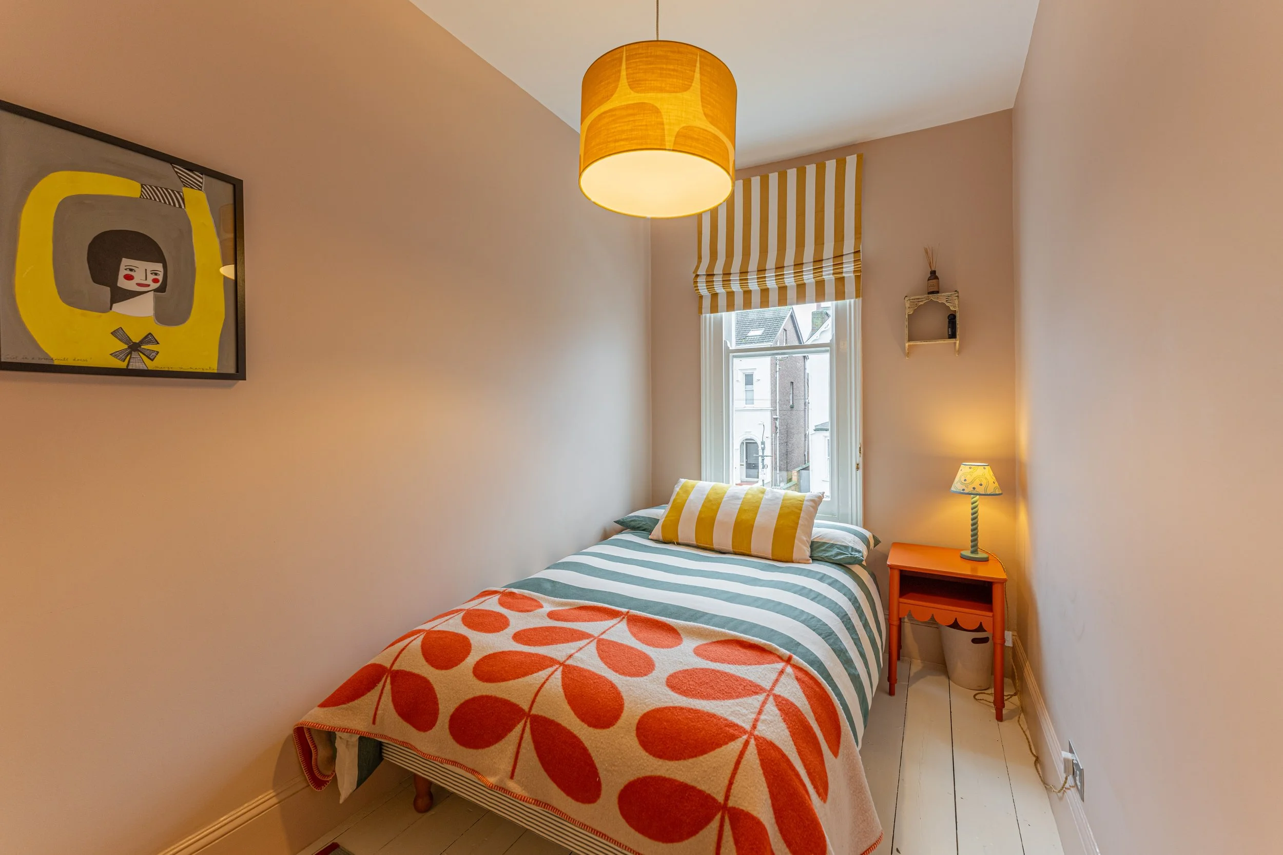 Small bedroom with striped bedding, a yellow-striped pillow, orange nightstand with a small lamp, window with striped window shade, wall art, and warm lighting