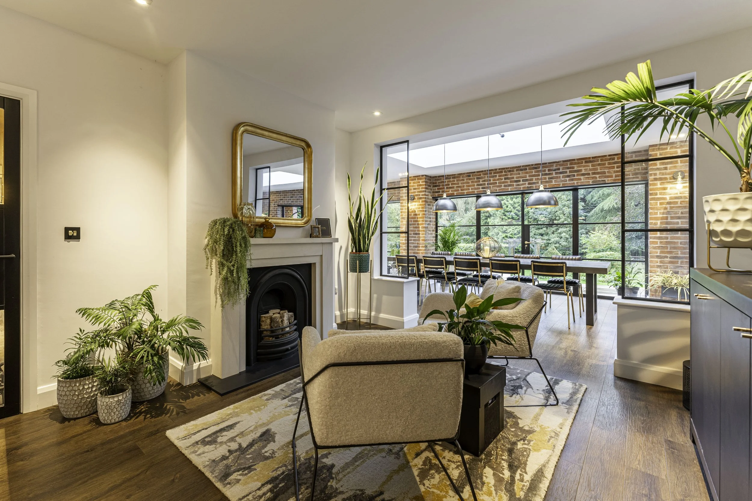 Modern living room with beige chairs, a white fireplace, and indoor plants, connected to a sunroom with a long dining table and large windows.