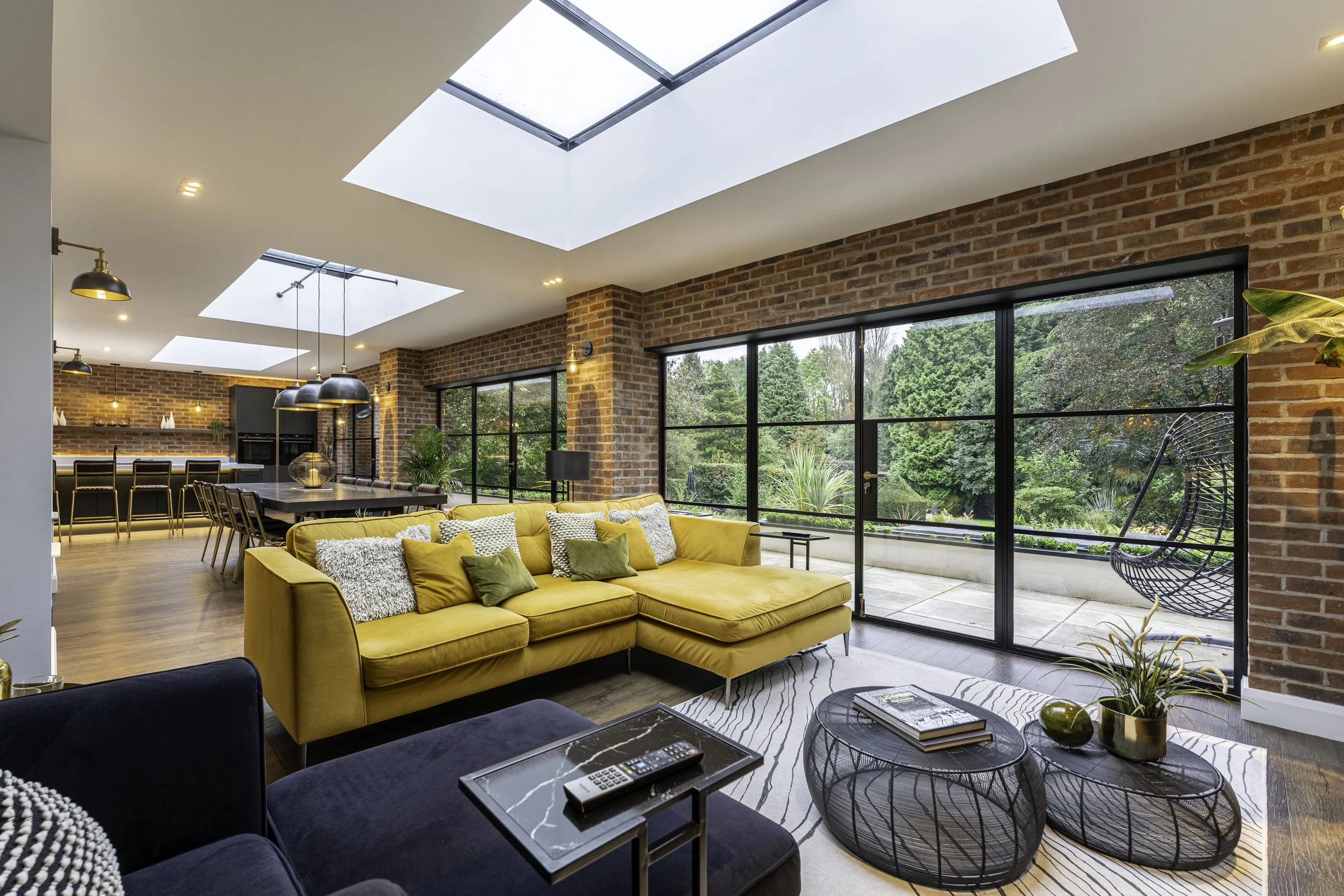 Open-concept living room with a yellow sectional sofa, black coffee tables, and large windows revealing a lush garden outside.