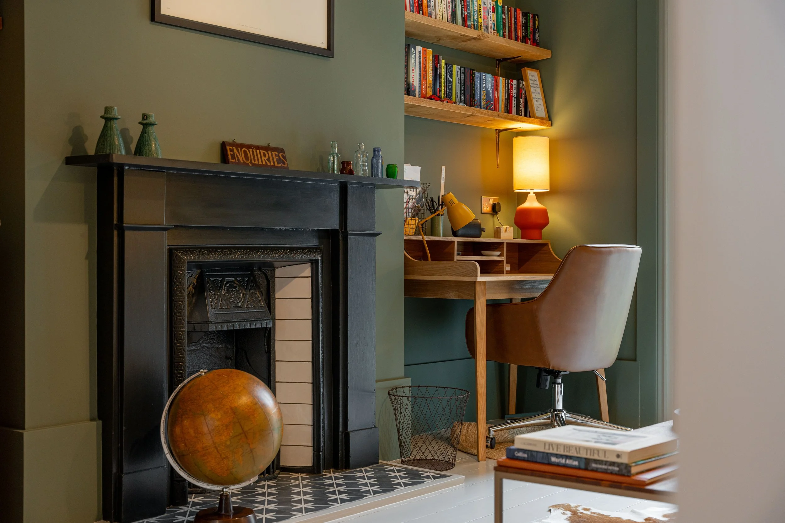 A cozy home office with a black fireplace, a globe, a wooden desk with a brown leather swivel chair, floating wooden shelves filled with books, a yellow lamp, and various decorative items on the mantel and desk.