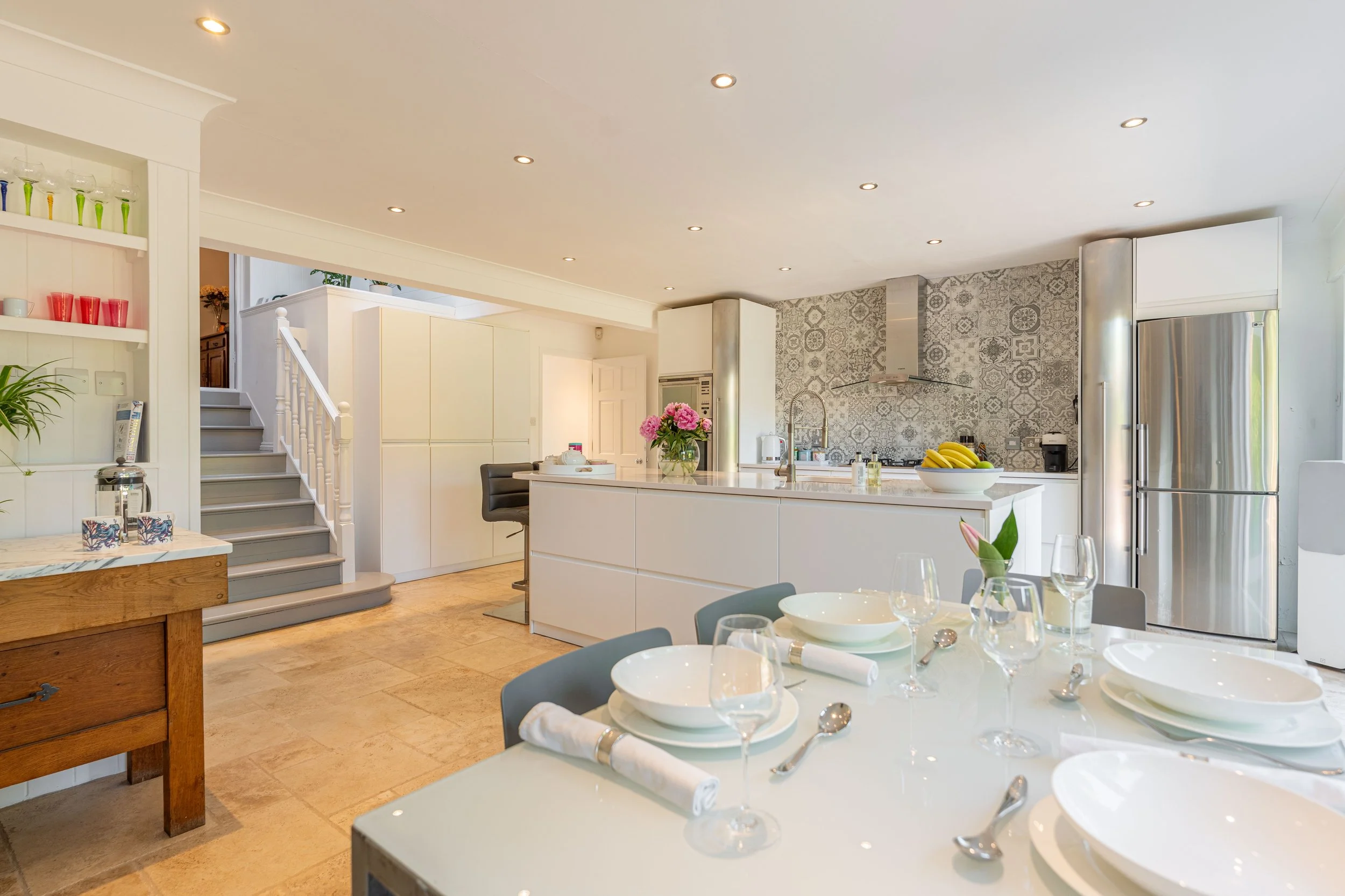 A spacious, modern kitchen with a white island counter, stainless steel refrigerator, patterned backsplash, dining table with place settings, and pink flowers in a vase.