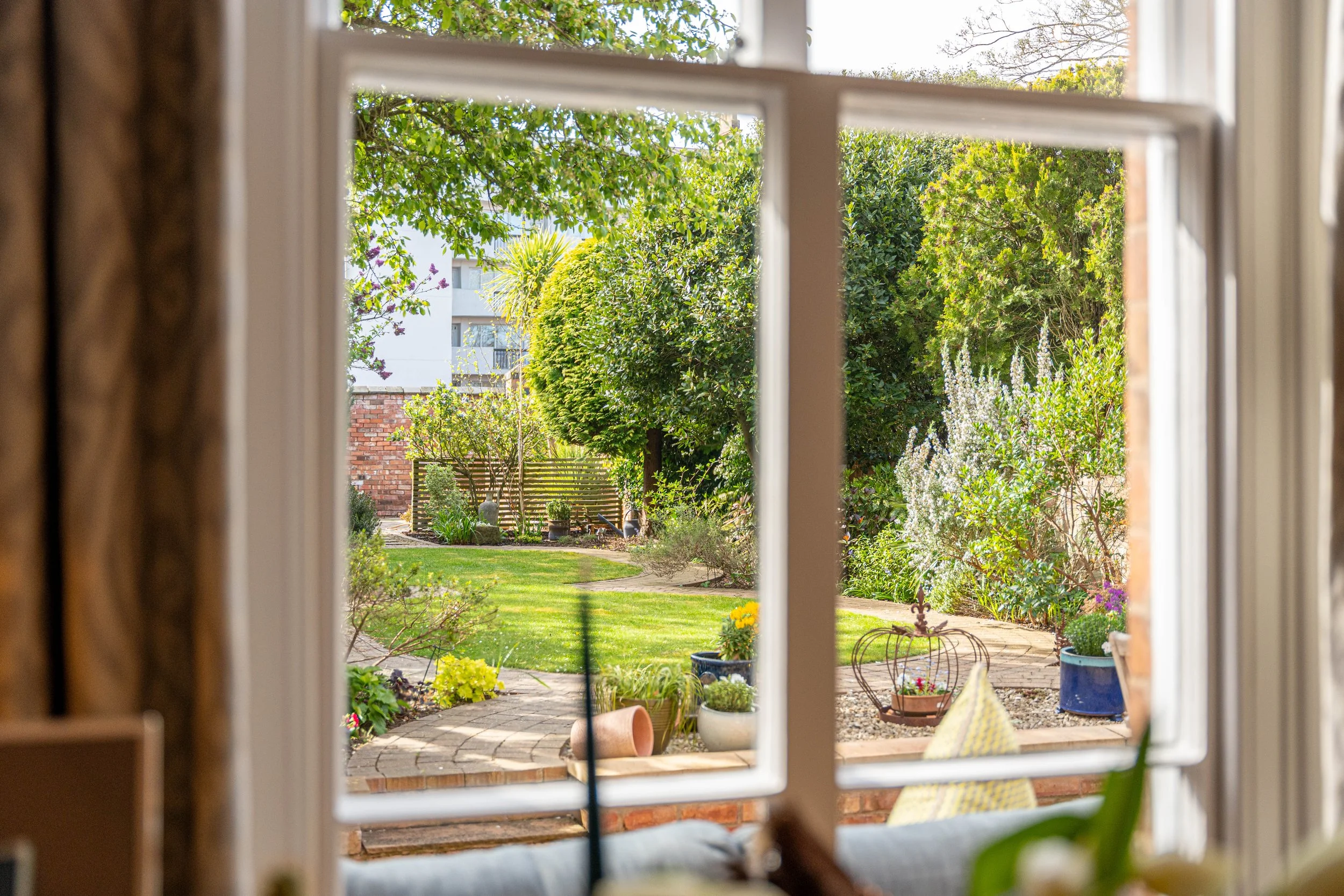 Garden view through a window with white frame, showing a lush yard with trees, bushes, a paved path, and potted plants.