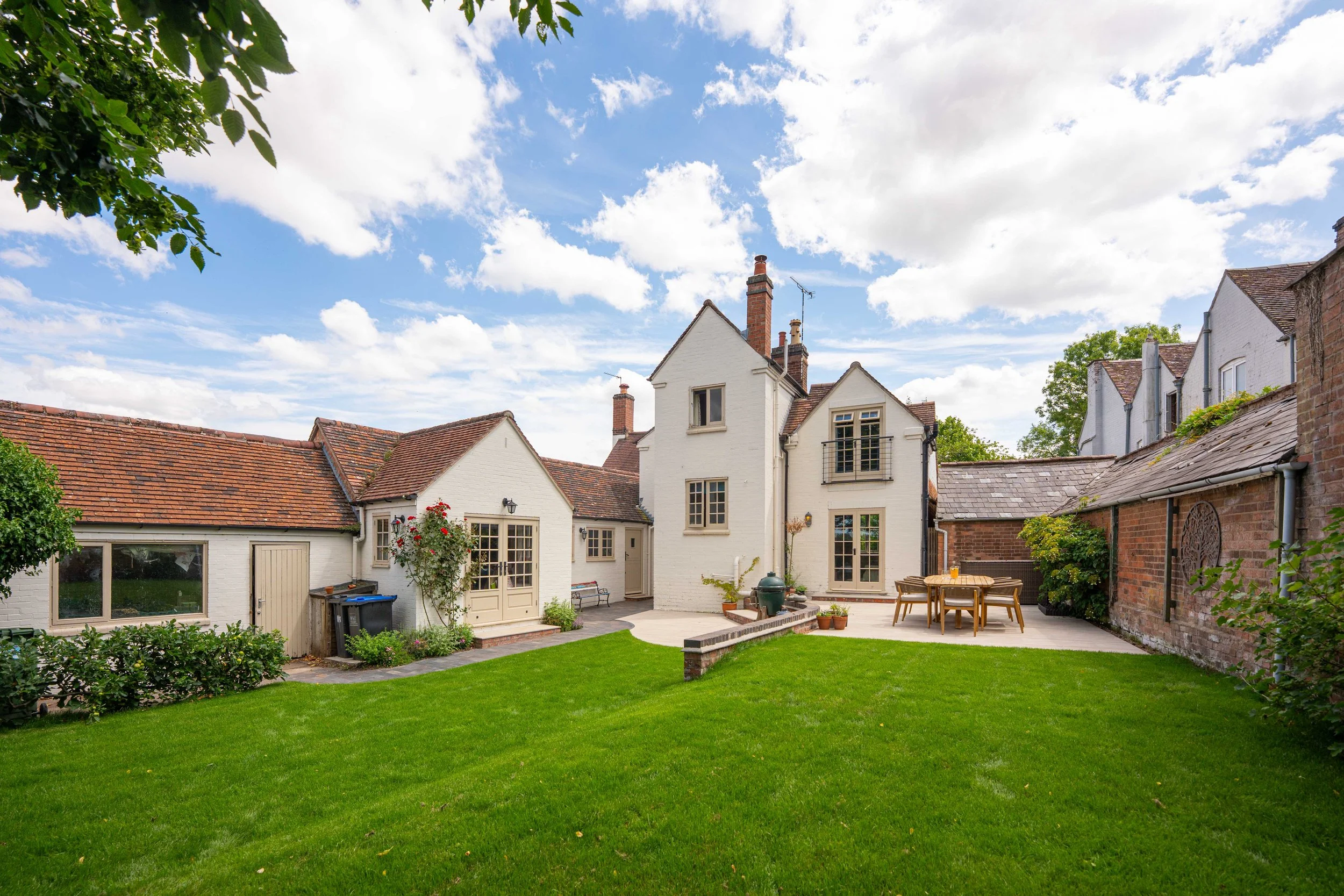 A backyard with a green lawn, a patio area with wooden furniture, and a white house with multiple windows and chimneys under a partly cloudy sky.