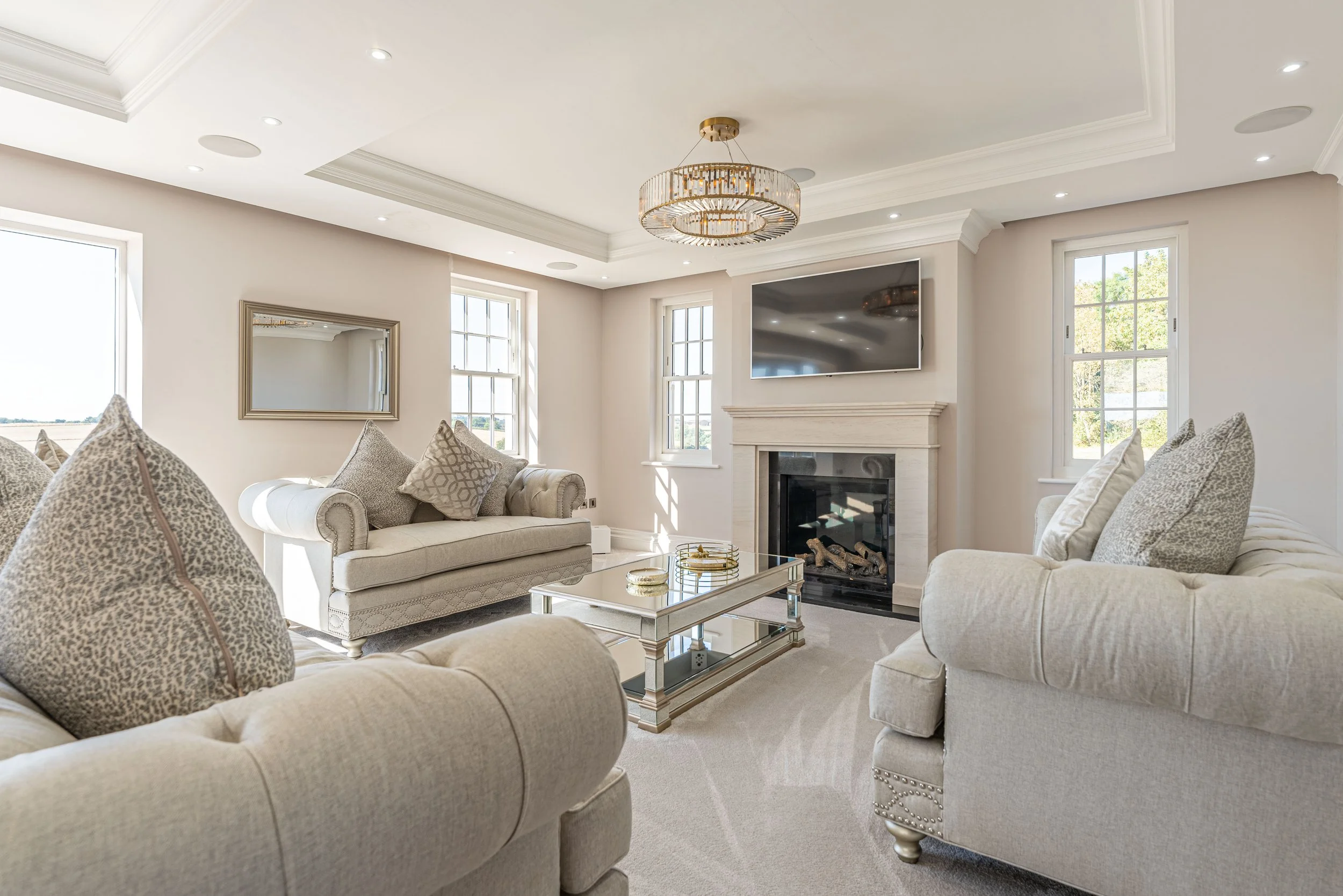 Elegant living room with cream-colored sofas, a glass coffee table, a fireplace, a wall-mounted TV, and large windows letting in natural light.
