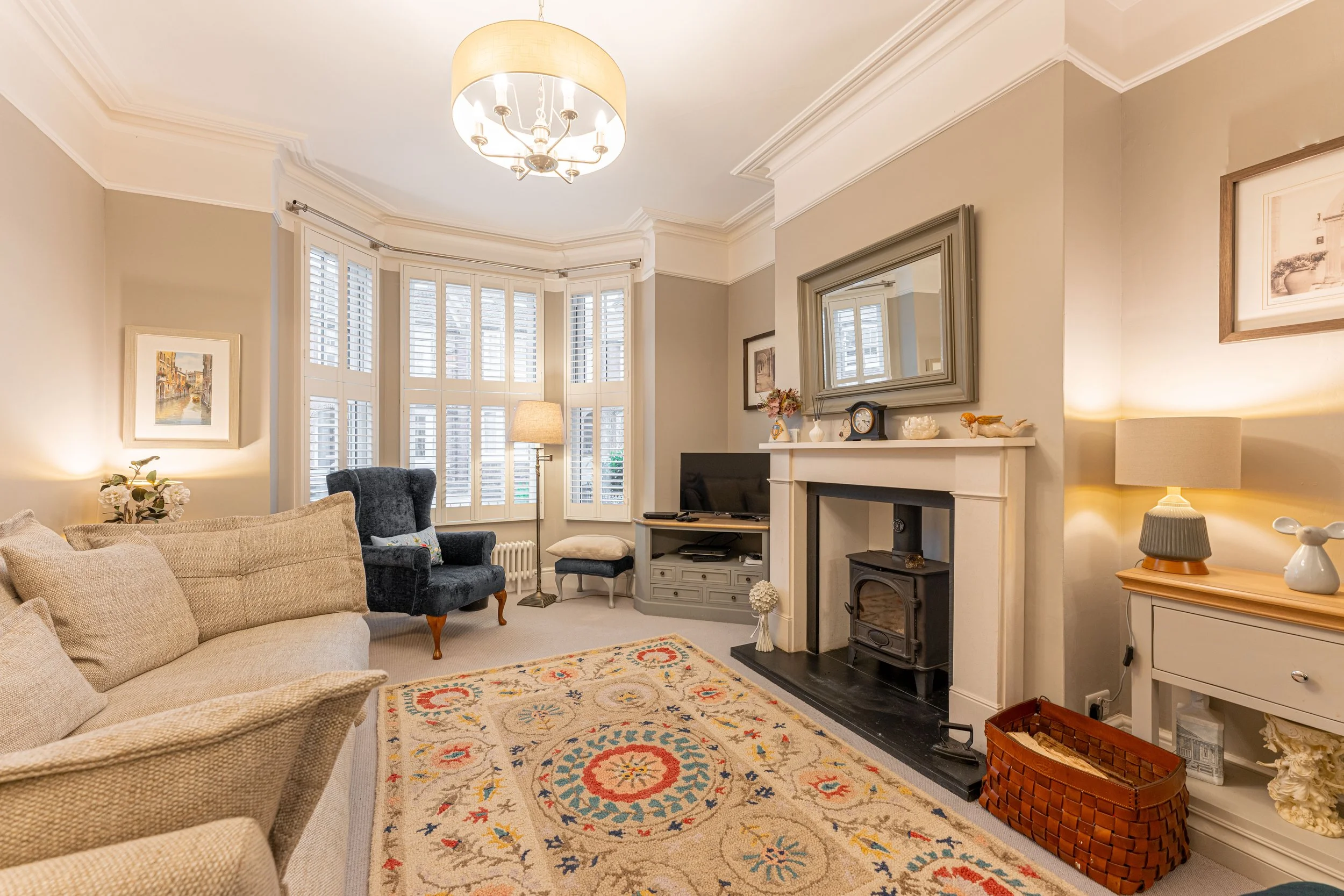 Cozy living room with beige sofa, navy armchair, window with white shutters, area rug, fireplace, TV, side table with lamp, and wall art.