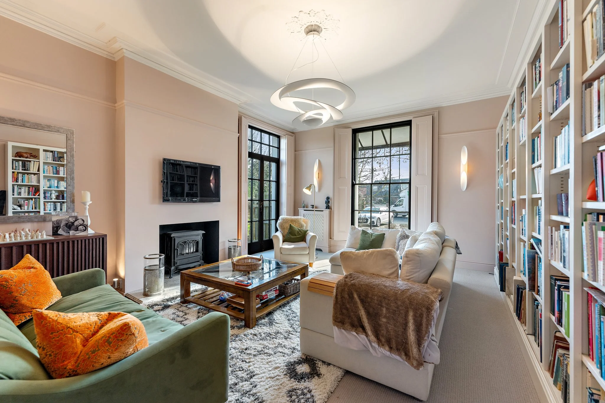 Living room with beige walls, large windows, a white sectional sofa with throws and cushions, a wooden coffee table, a TV above a fireplace, bookshelves filled with books, a mirror, and decorative lighting fixtures.
