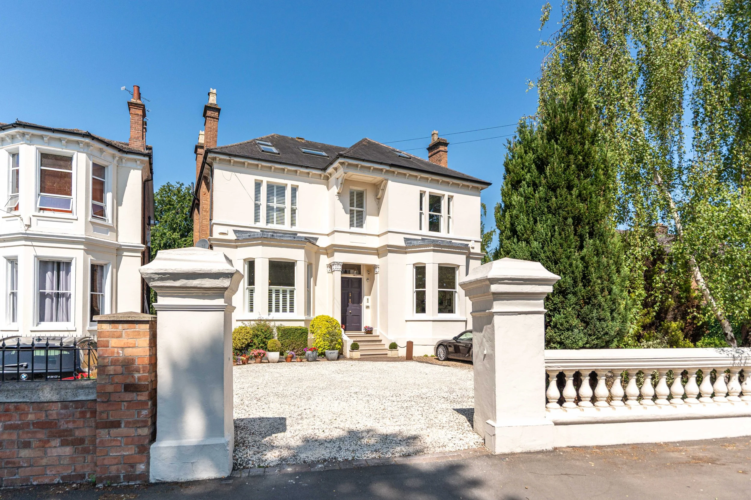 White two-story house with black front door, gravel driveway, and potted plants in front; tall trees and a blue sky in background.