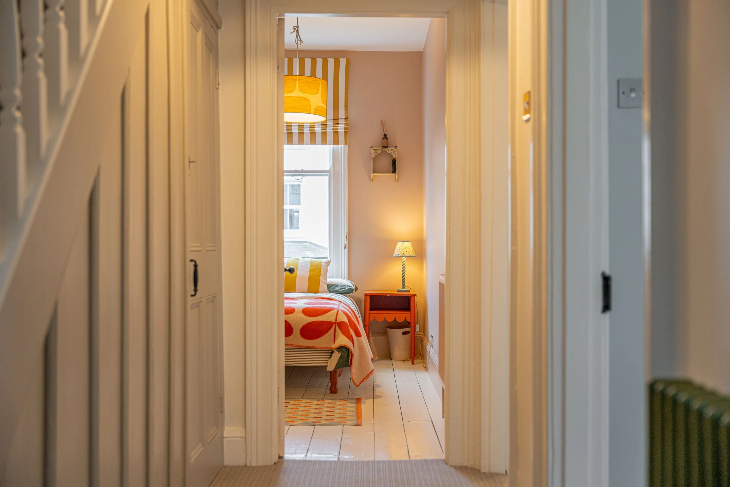 View through a hallway into a cozy bedroom with a bed, a side table with a lamp, a window with striped curtains, and decorative wall shelves.