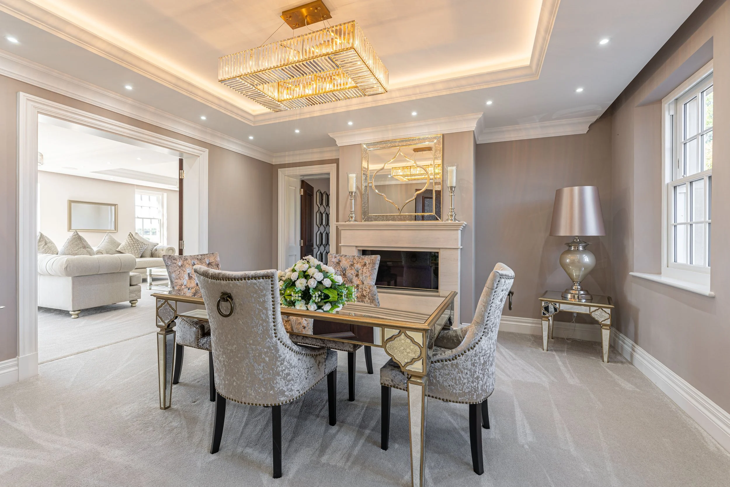 Elegant dining room with a rectangular table, six upholstered chairs, a floral centerpiece, a fireplace, a large mirror above the fireplace, a chandelier, and a side table with a lamp, with a living room visible through an open doorway.