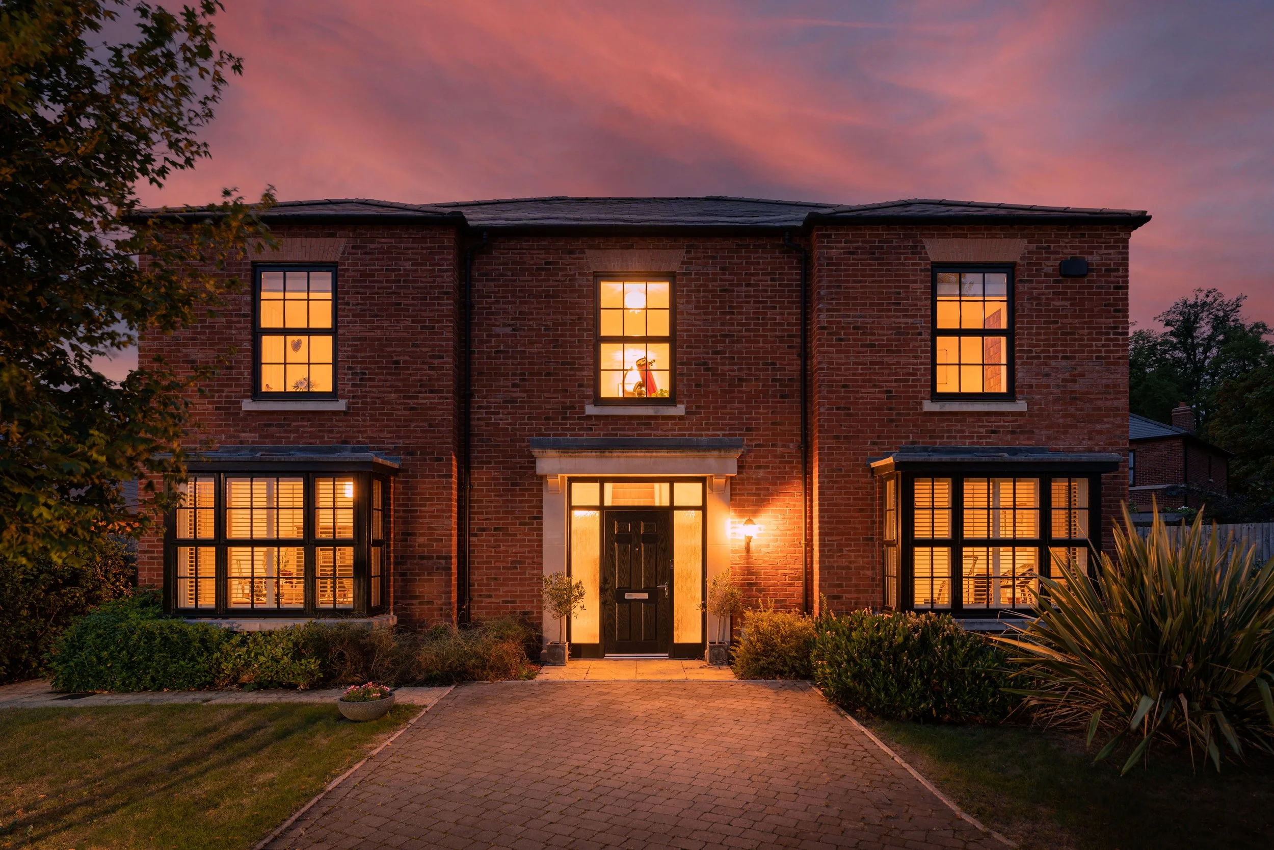 A brick house at sunset with warm lights in the windows, a paved pathway leading to the front door, surrounded by greenery and a colorful sky.