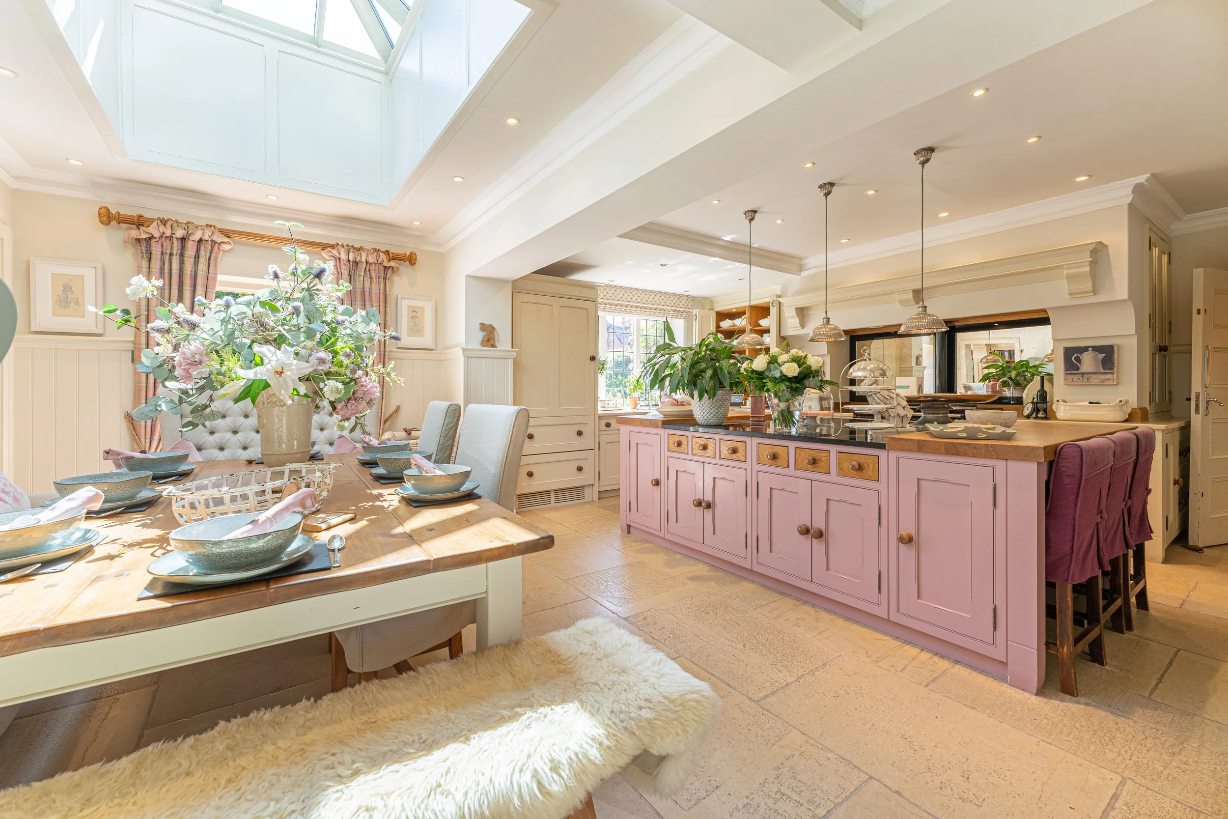 A bright and airy kitchen and dining area with a large skylight and sunlight streaming in. There is a wooden dining table set with blue dishes and pink napkins, a fluffy white bench, and a pink kitchen island with a wooden countertop. The space is decorated with potted plants and floral arrangements.