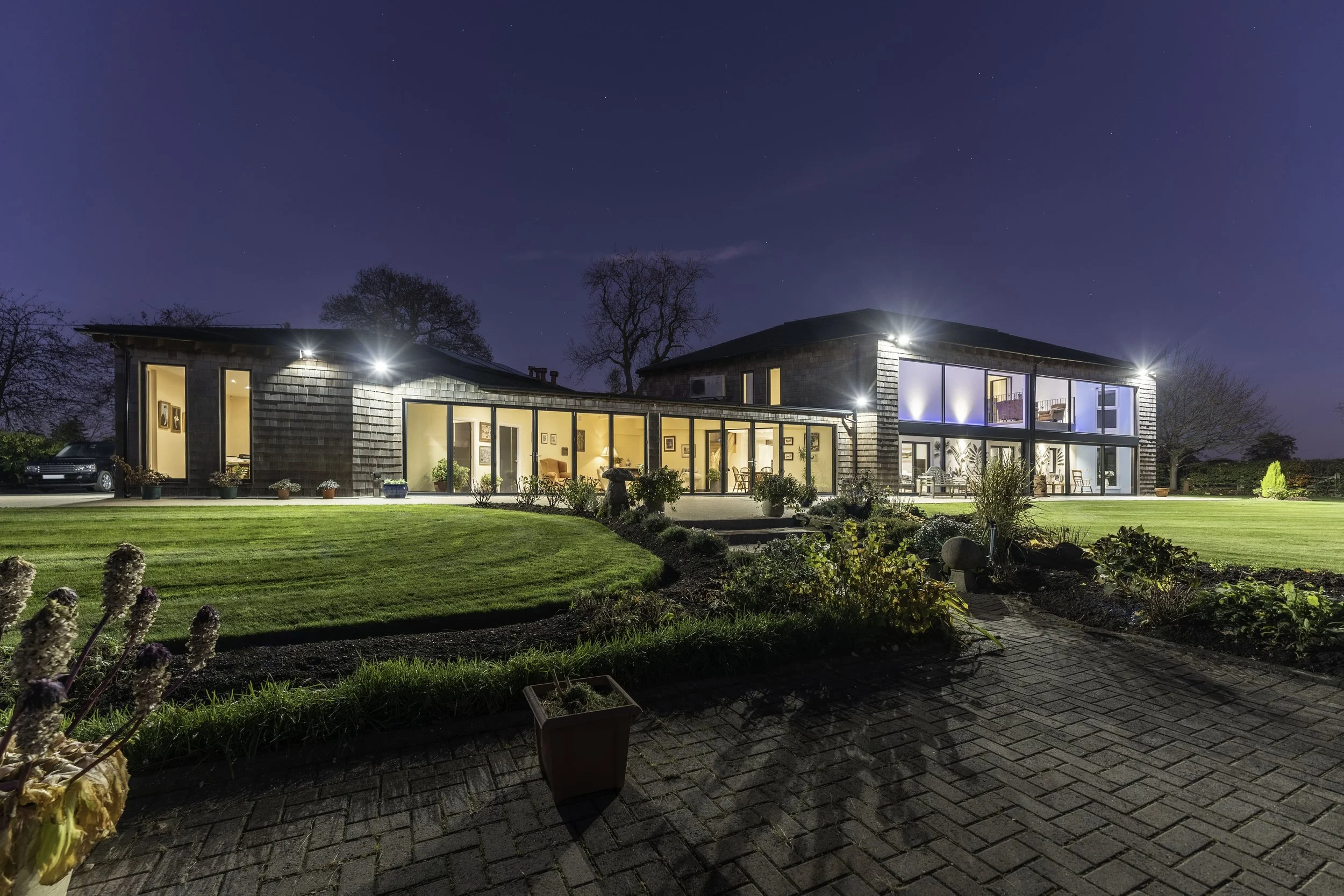 Modern house illuminated at night, with large glass windows, a well-kept lawn, and garden, under a starry sky.