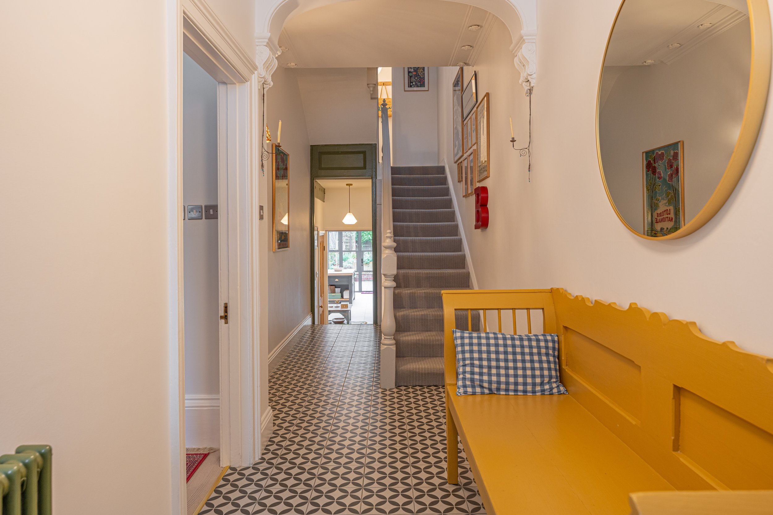 Interior view of a house hallway featuring a patterned tile floor, a yellow wooden bench with a blue and white gingham pillow, a staircase with carpeted steps, framed pictures on the wall, a large round mirror, and a view towards the living area with
