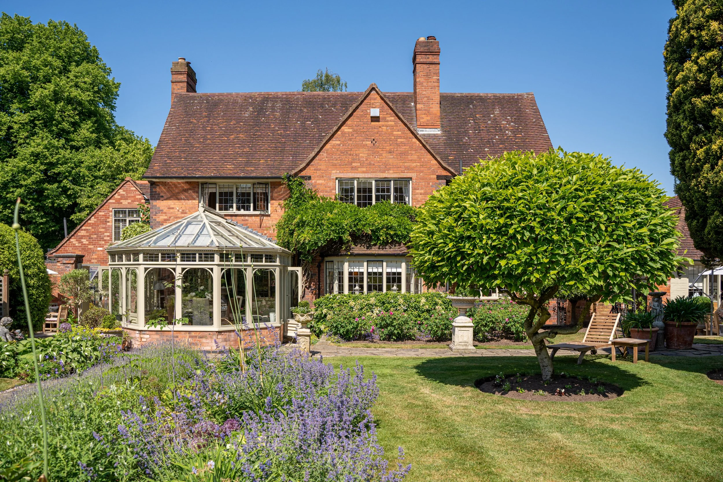 A large brick house with a triangular roof, surrounded by a lush garden featuring colorful flowers, a neatly mowed lawn, and a tree with dense green foliage. There is a glass conservatory attached to the house on the left side and garden benches.