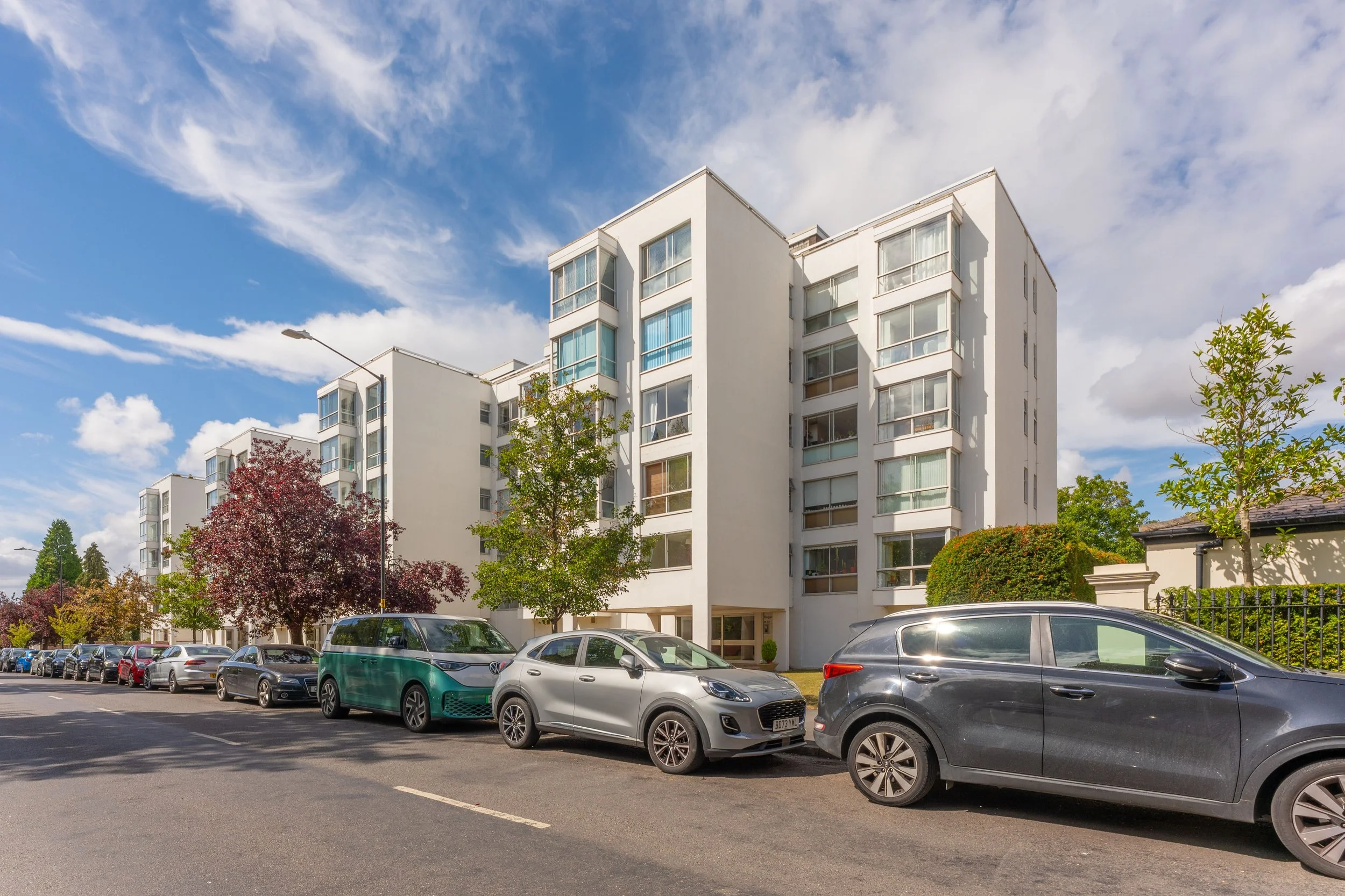 Modern white apartment building with multiple stories, large windows, and balconies, alongside parked cars and trees under a blue sky with clouds.