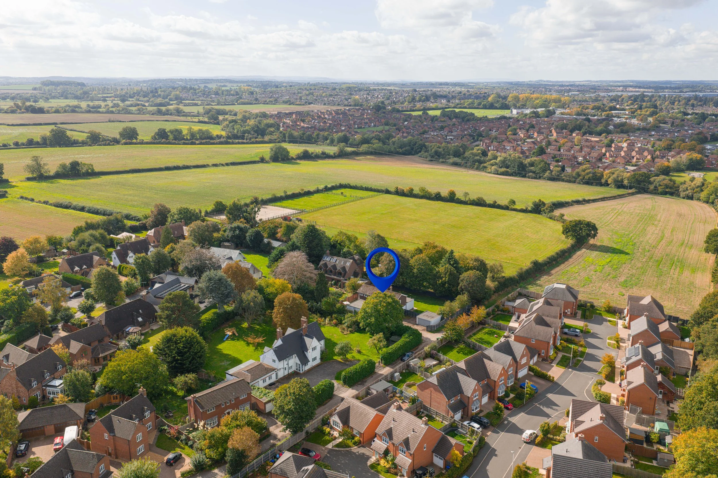 Aerial view of a residential neighborhood with houses, greenery, and large open fields in the background under a partly cloudy sky.