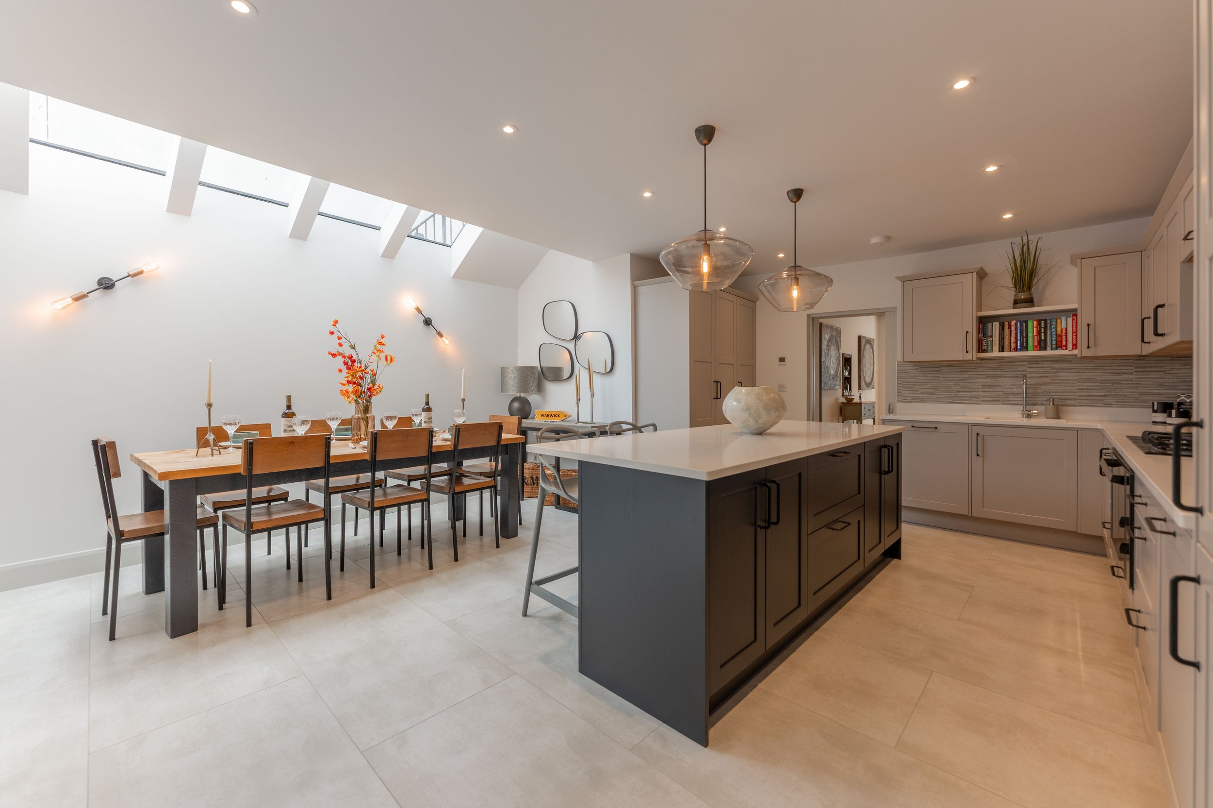 Open-concept kitchen and dining area with skylights, modern lighting, beige cabinets, black kitchen island, and wooden dining table with nine chairs.