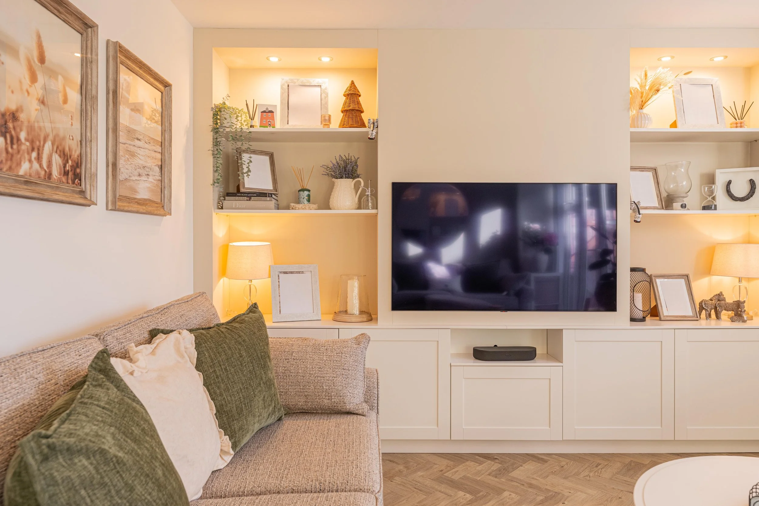 Living room with beige couch, green and white pillows, wall-mounted TV, and built-in shelves decorated with framed photos, vases, and books. Two lamps with beige shades provide warm lighting.