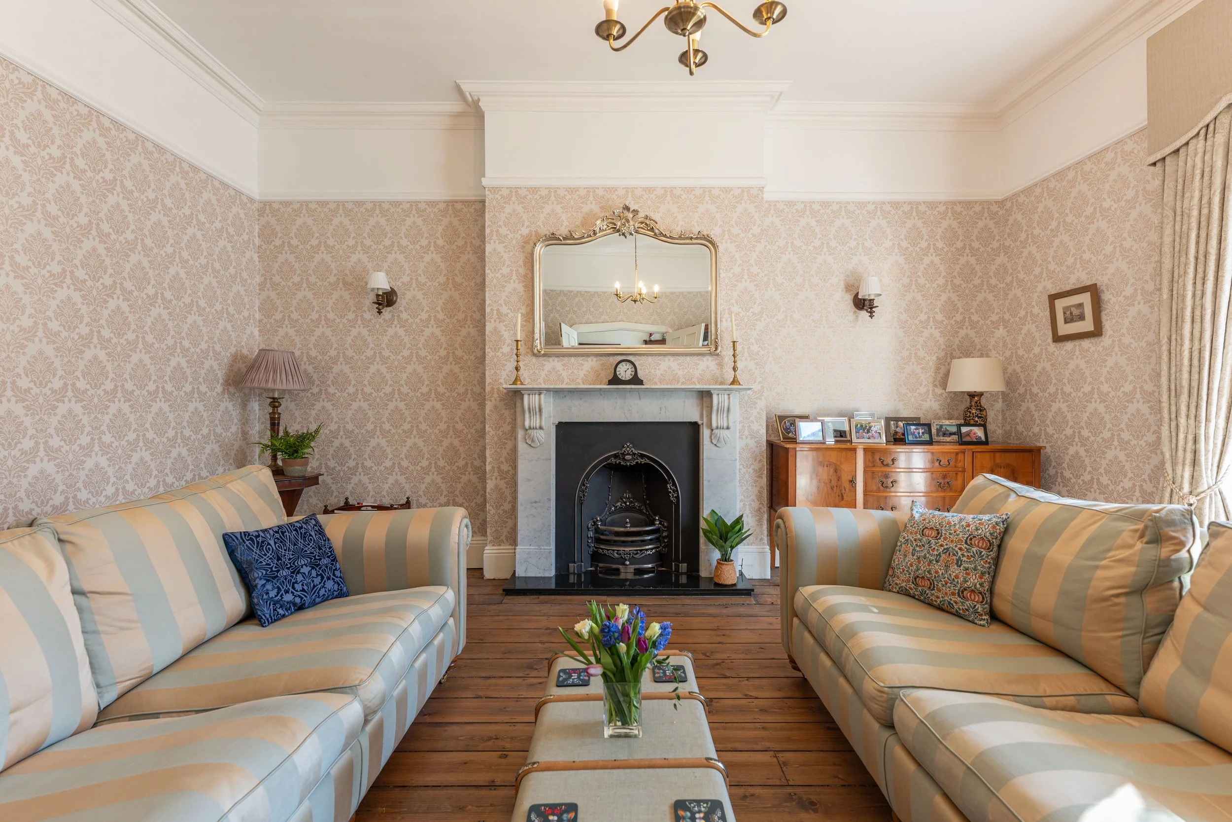 Living room with two striped sofas, a fireplace with a mirror above it, wooden floors, floral wallpaper, and side tables with lamps and framed photos.